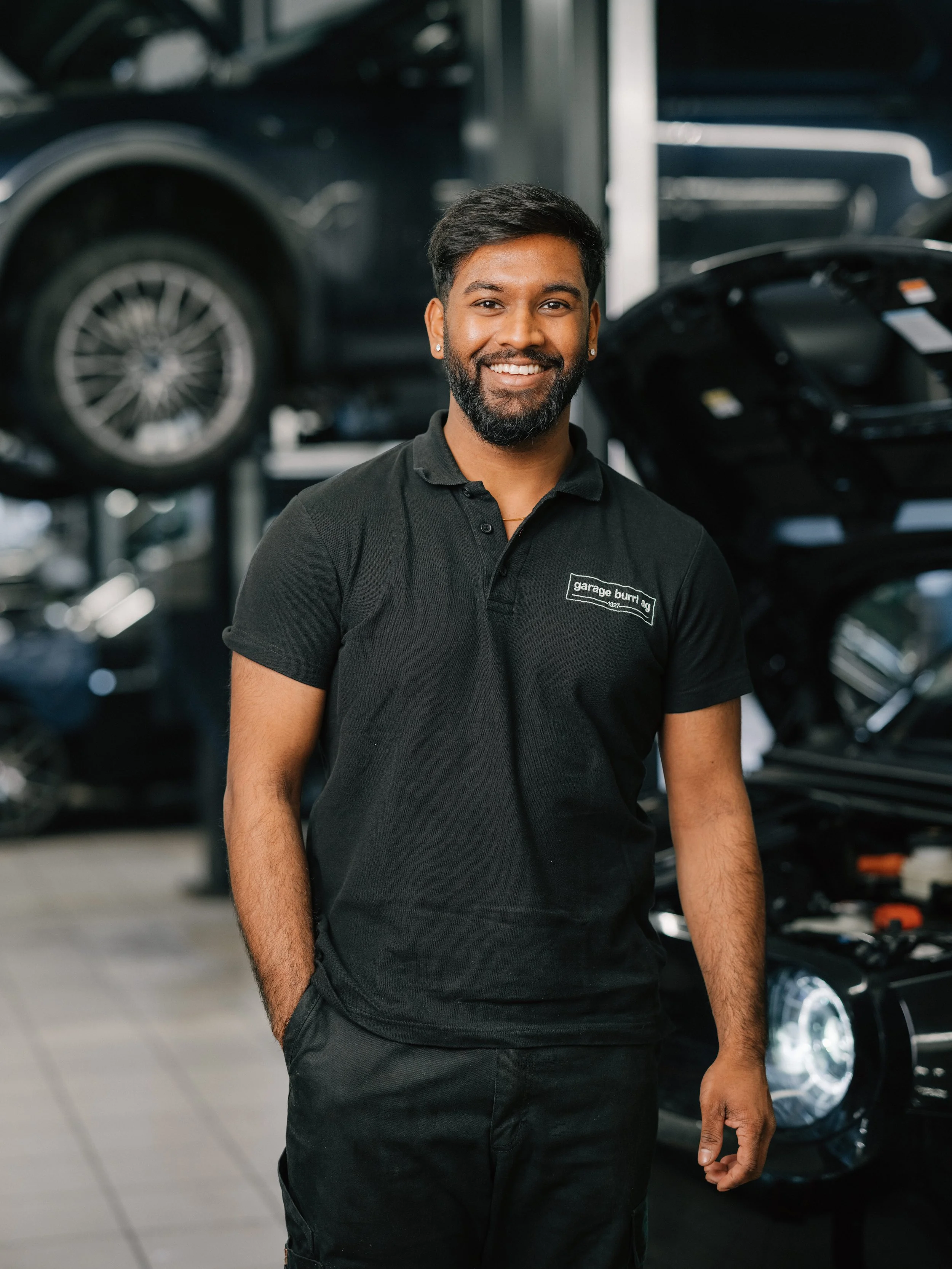 Lächelnder Mann mit dunkler Haut, schwarzem Haar, Bart und weißem Hemd vor einer weißen, modernen Wand.