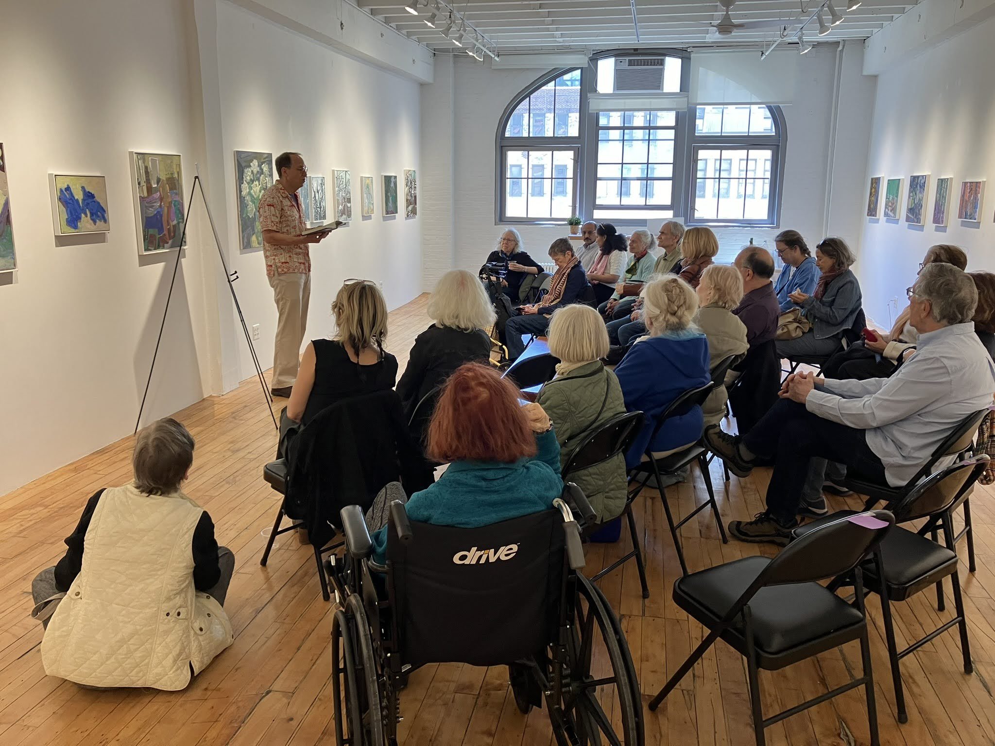 A man giving a presentation or lecture to an audience seated in a gallery or art space with paintings on the walls and large windows in the background.