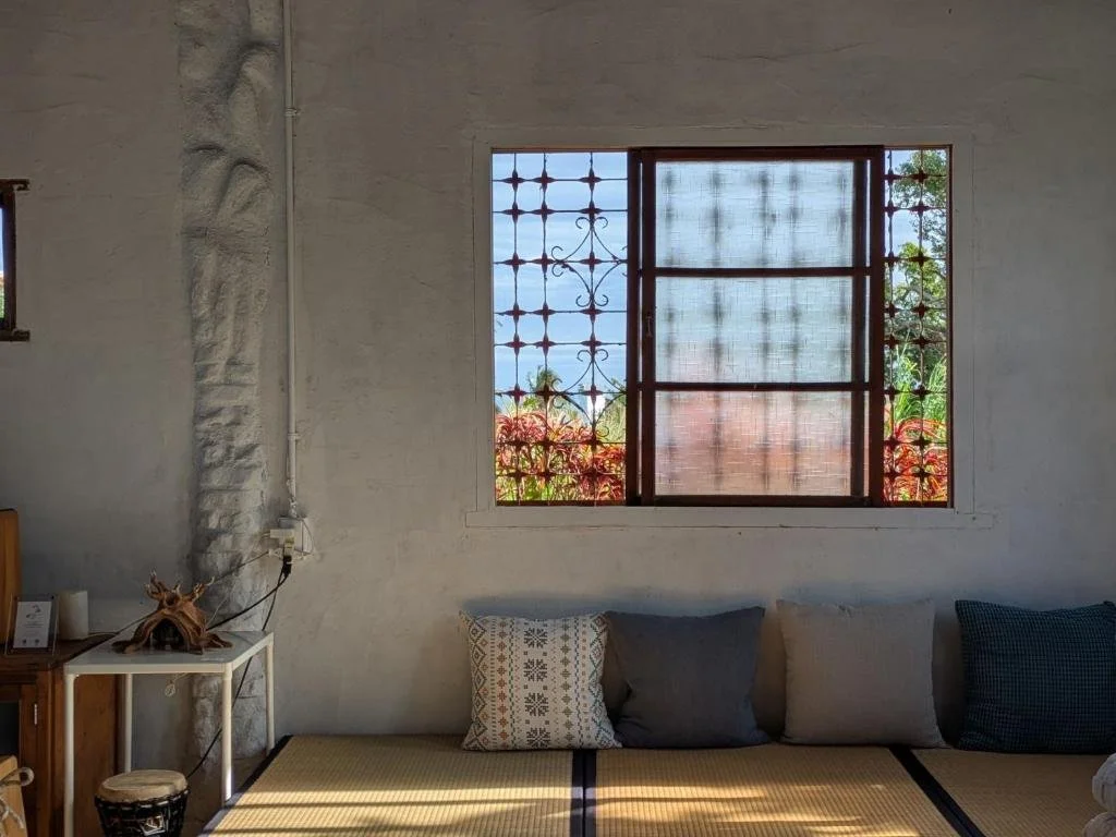 A room with a window showing a metal security grille and a view of bushes and trees outside. The room has a sofa with four cushions, a small table with decorative items on it, and a textured white wall.