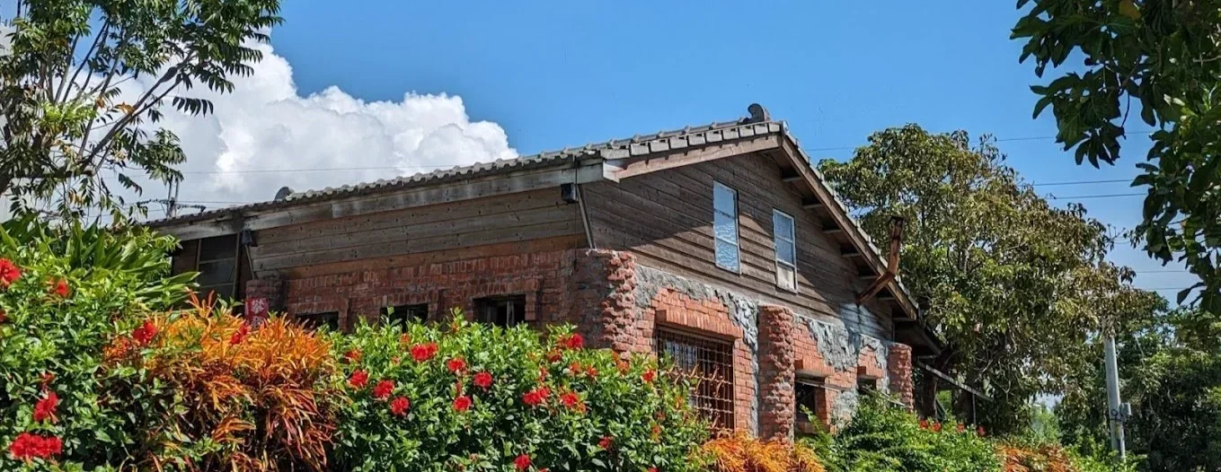 A rustic house with brick and wood construction, surrounded by colorful flowers and trees under a bright blue sky with white clouds.