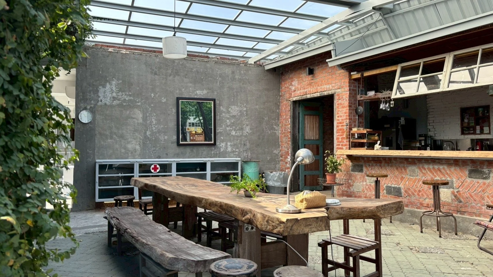 Indoor patio with rustic wooden dining table, benches, and stools. Surrounded by brick and concrete walls, with plants, a clock, and kitchen visible in the background. Glass roof overhead.