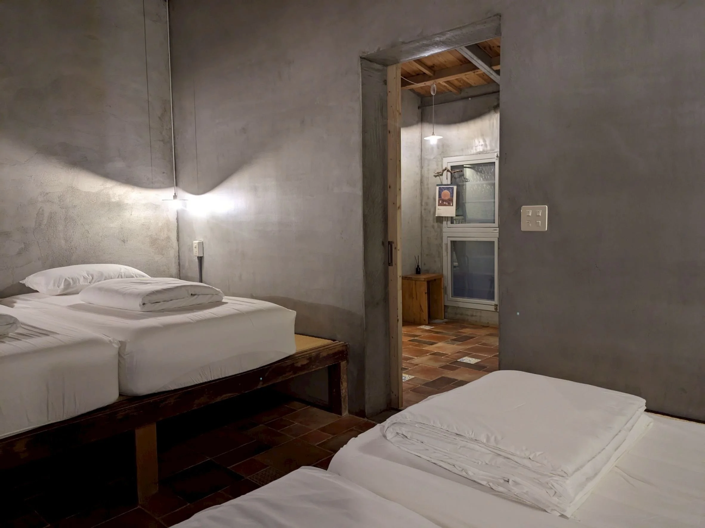 Two beds with white linens in a minimalist bedroom with exposed gray concrete walls and brick flooring, doorway leading to a small room with window, wooden ceiling, and hanging light.
