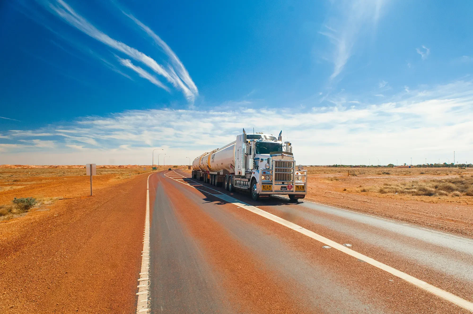 semi trailer truck driving along a highway. There are dry, baron fields on either side of the highway comprised of a lot of dirt and some spots of grasses