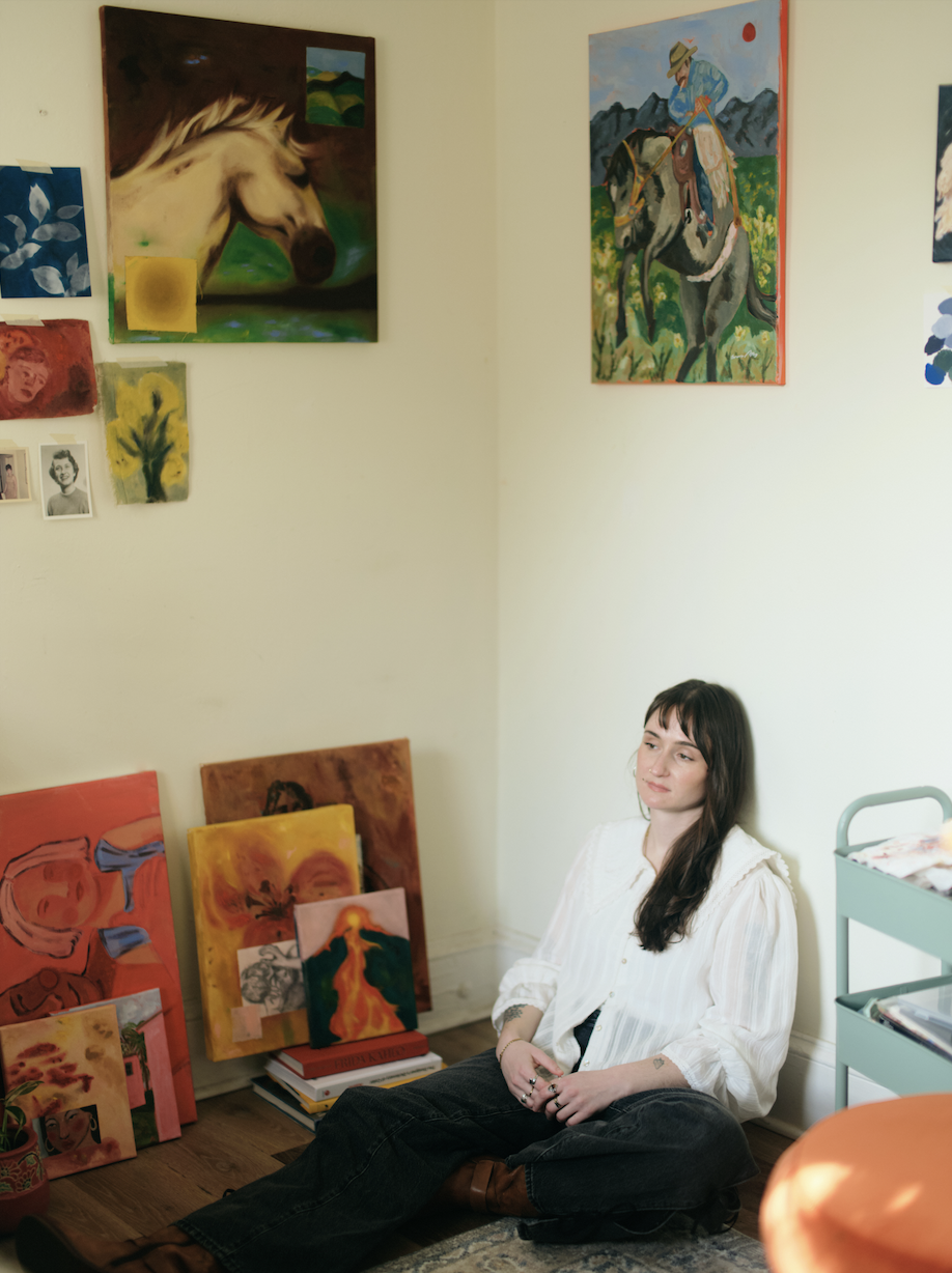 A woman sitting on the floor against a wall decorated with various colorful paintings, including horse and mountain scenes, in an art studio or gallery.