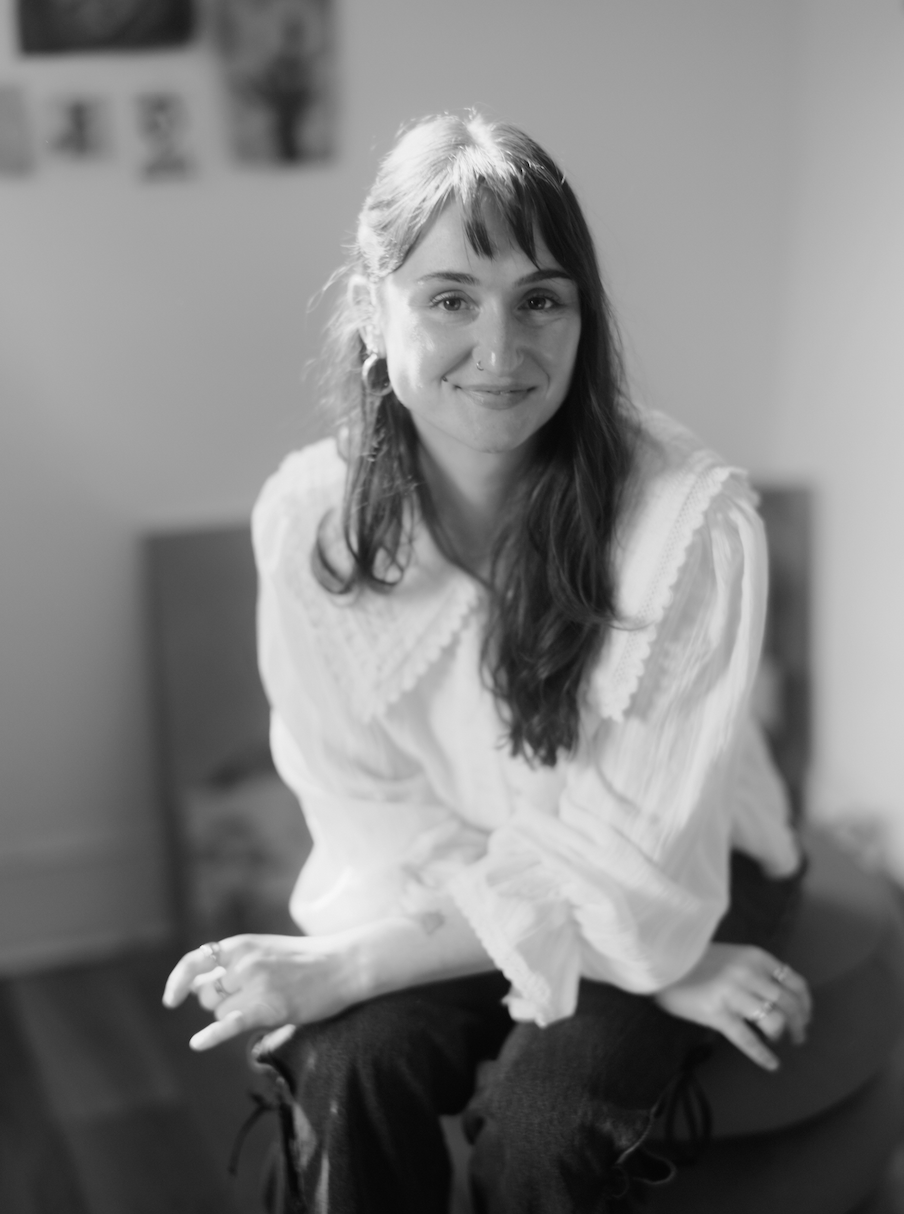 A woman with long hair and a nose piercing sitting on a bench, smiling at the camera in a room with a blurred background.