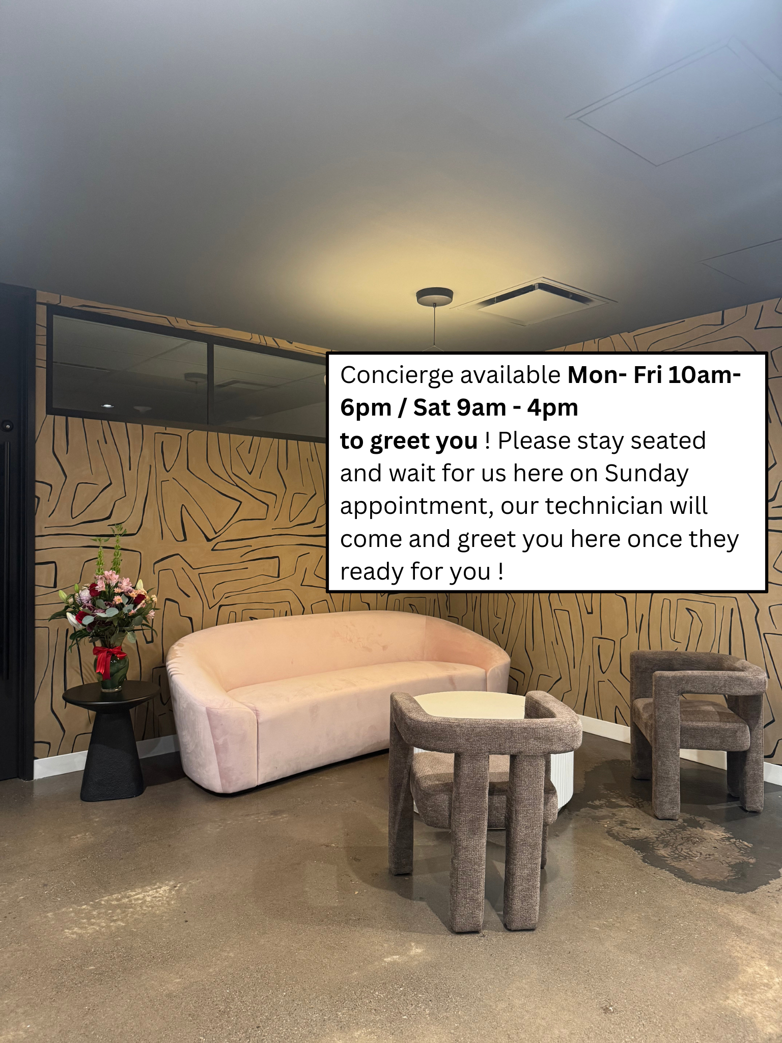 A waiting area with a pink sofa, two grey armchairs, a black side table with a bouquet of flowers, and a wall with modern geometric patterns. A sign with appointment details and available concierge hours is partially visible.