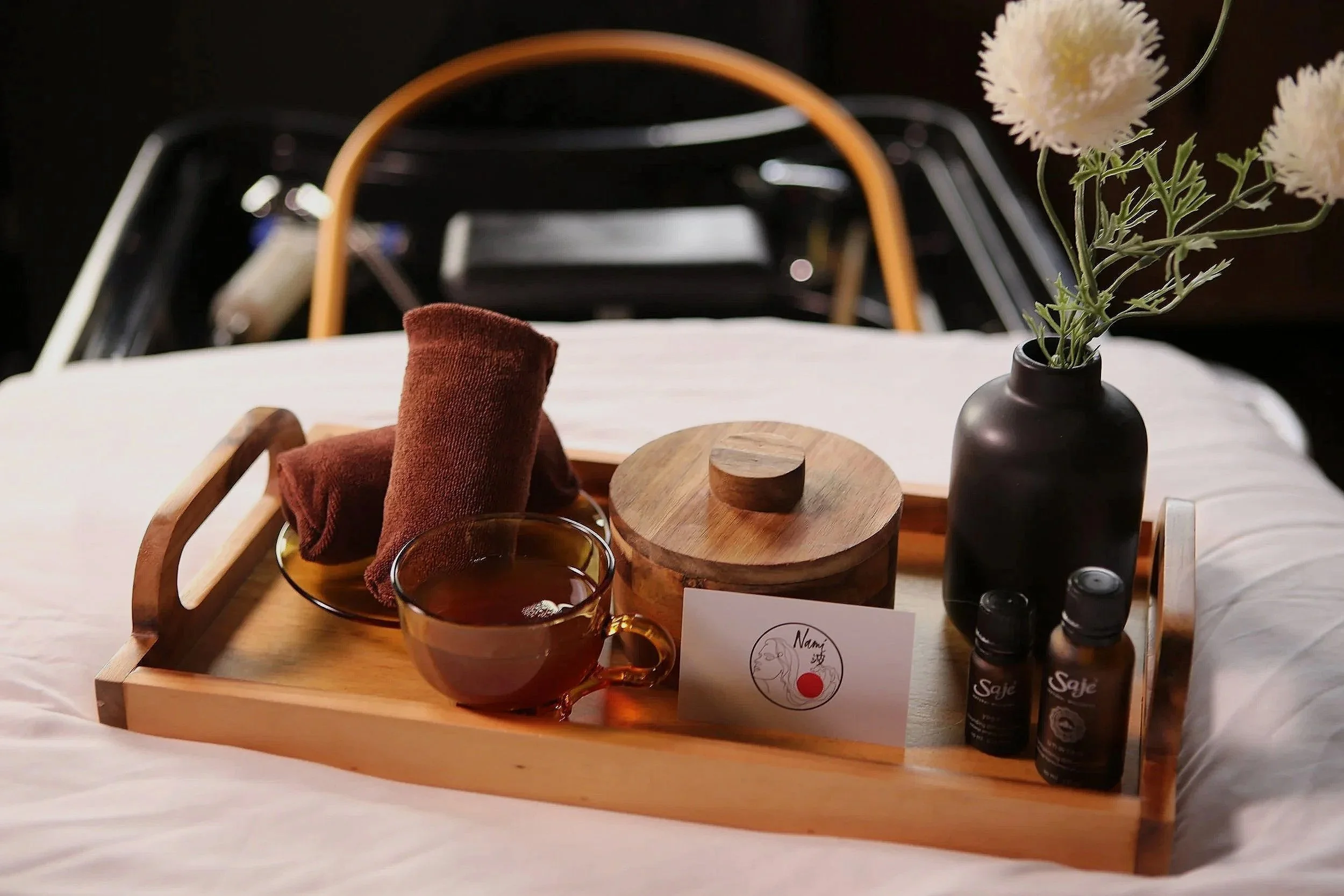 A wooden tray holding two glasses of tea, a small round wooden box with a lid, a small black vase with white flowers, and bottles of scented oils. The tray is on a bed with white bedding, with a headboard and a black background visible behind.