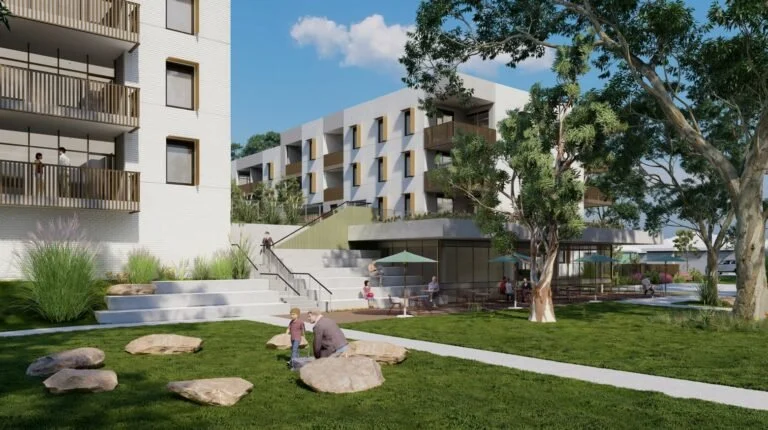 Modern residential apartment complex with outdoor communal space, trees, green grass, rocks, and people sitting and walking outside.