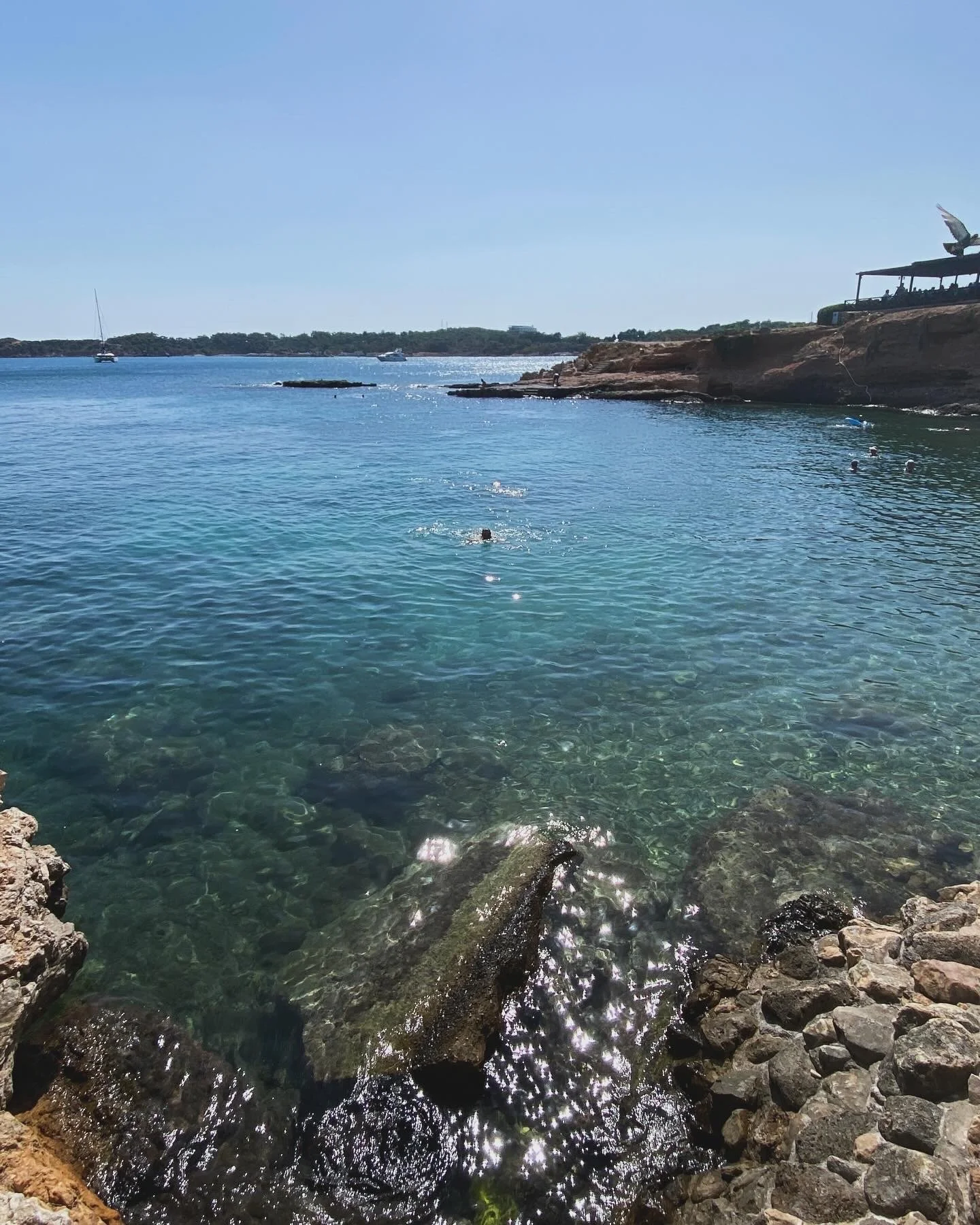 People swimming in clear blue water near rocky shoreline under a sunny sky, with boats and a structure on the rocky cliff in the background.