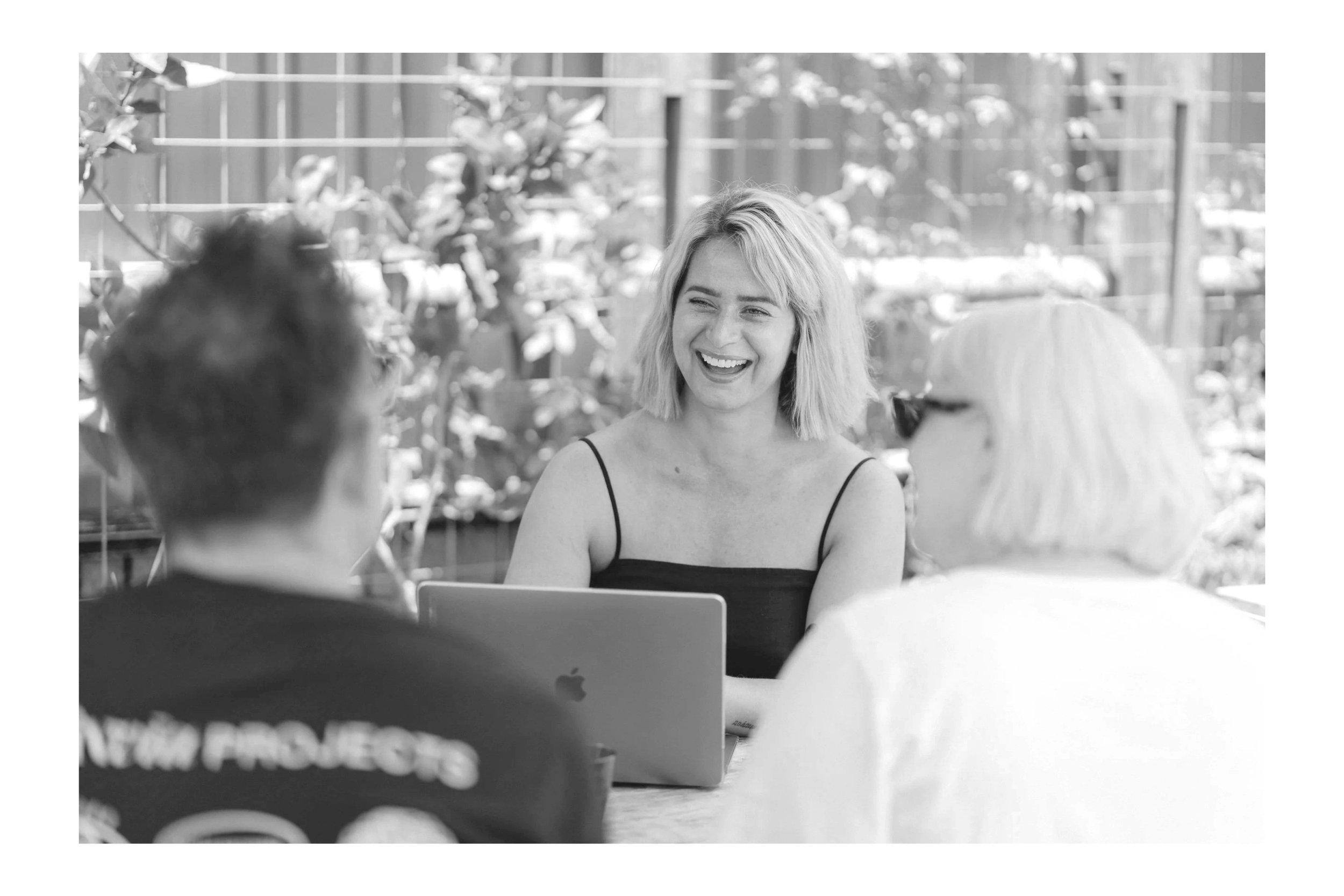 Two women and one man sitting together outdoors, smiling and talking, with one woman in the center having a laptop in front of her.