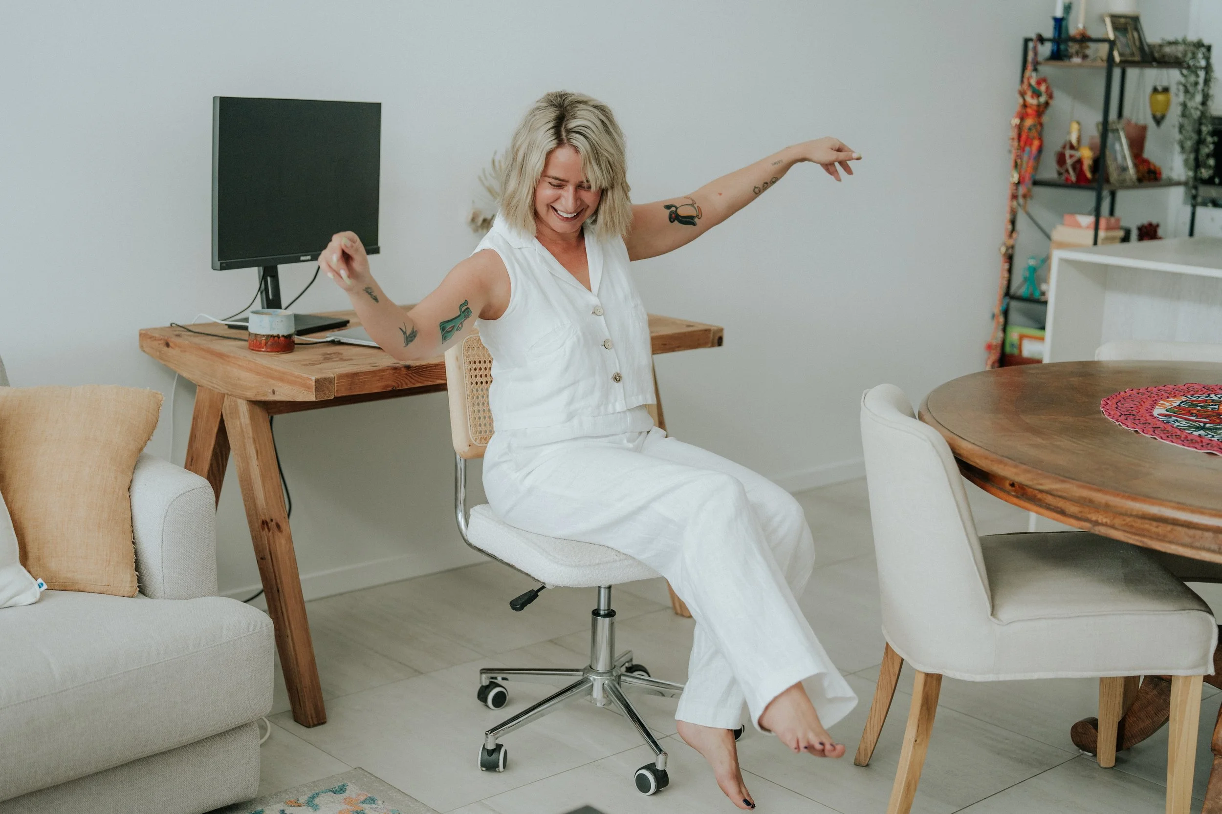 Woman with short blonde hair, wearing a white sleeveless top and pants, sitting on an office chair, smiling and dancing in a living room with a wooden desk, a computer monitor, and decorative shelves.