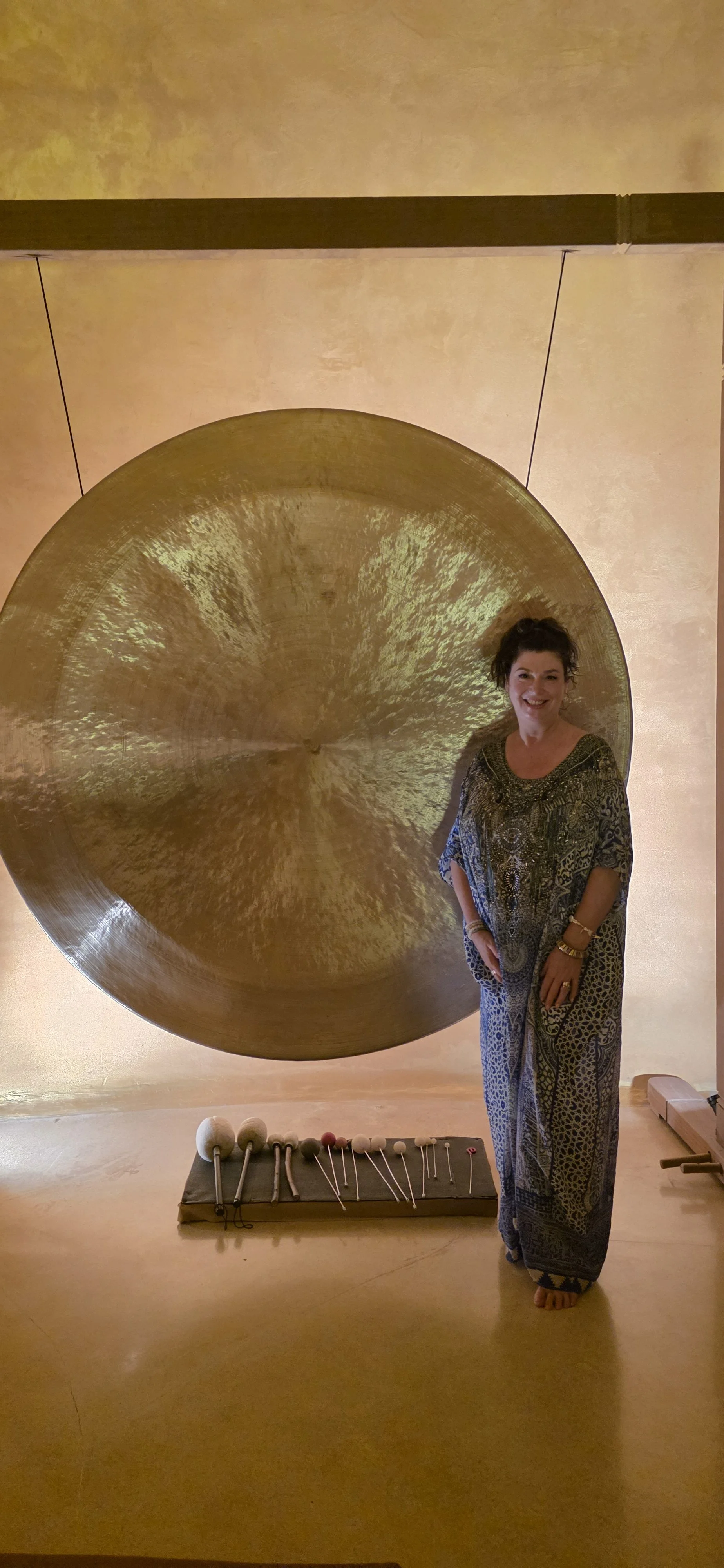 A woman standing next to a large hanging gong in a room with warm lighting. In front of her, there is a platform with various mallets and sticks used for playing the gong.