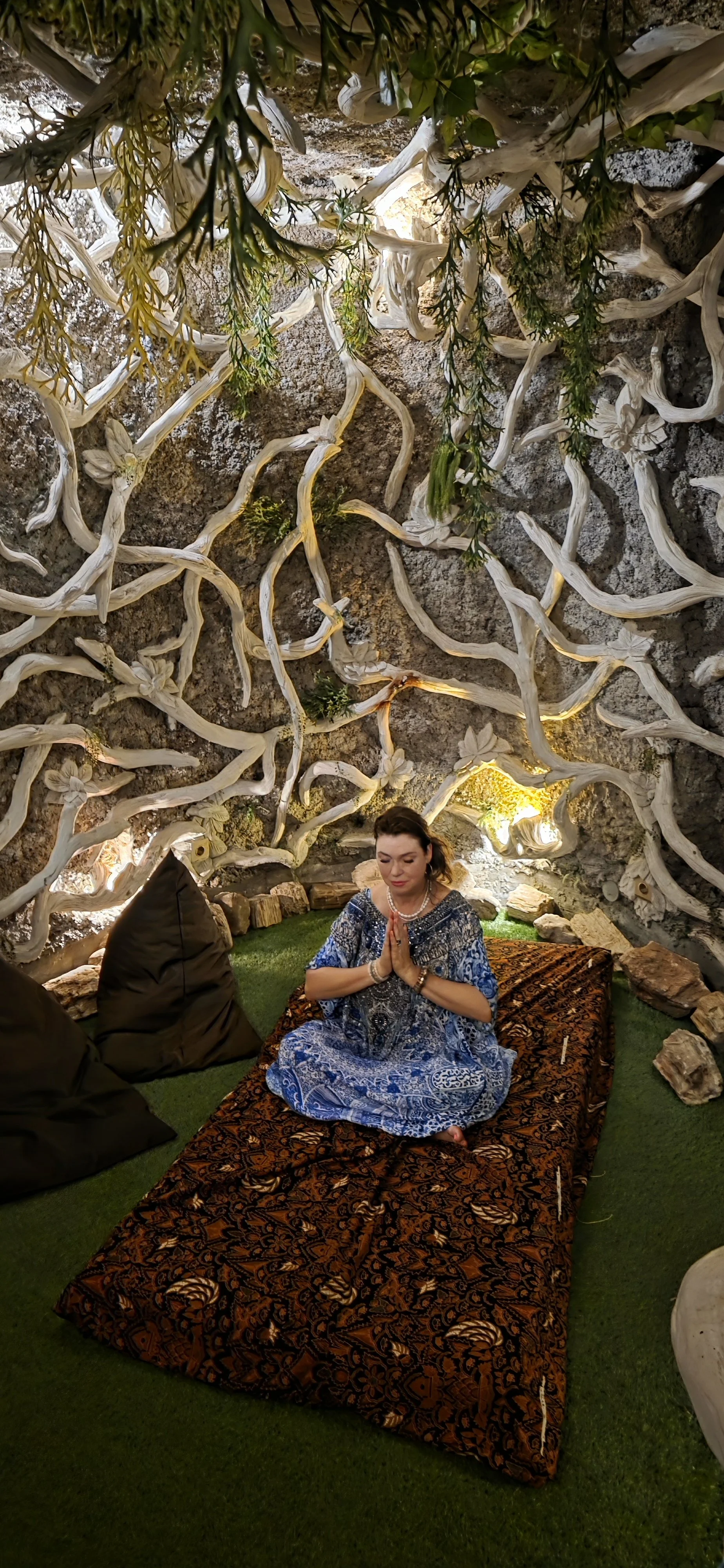 A woman dressed in blue, sitting on a patterned mat with her hands in prayer in a cave-like setting decorated with intertwined white branches and rocks, with soft lighting.