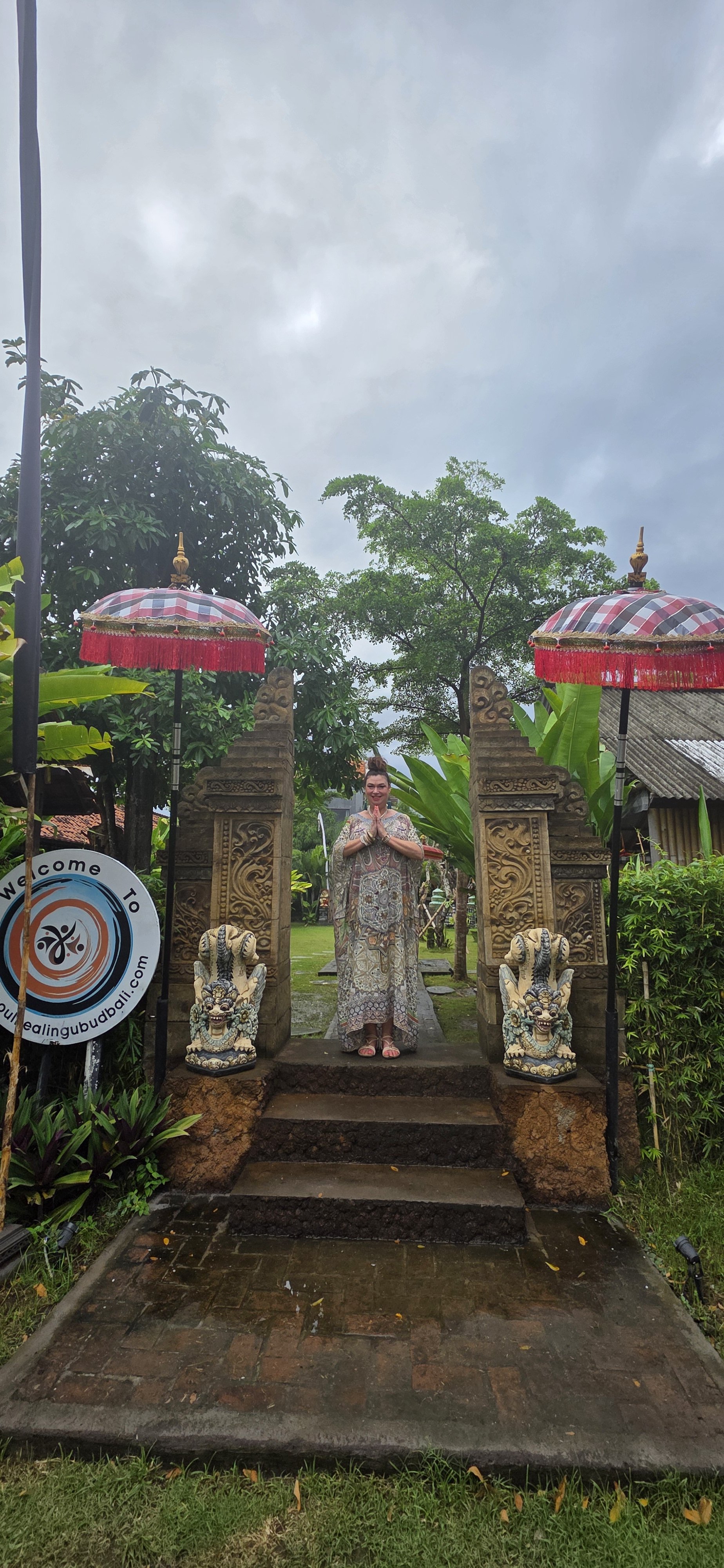 A woman standing with hands in a prayer gesture, surrounded by traditional Balinese elements including ornate stone statues, carved gateways, and umbrellas with red fringe, set in a lush garden with trees and a cloudy sky.