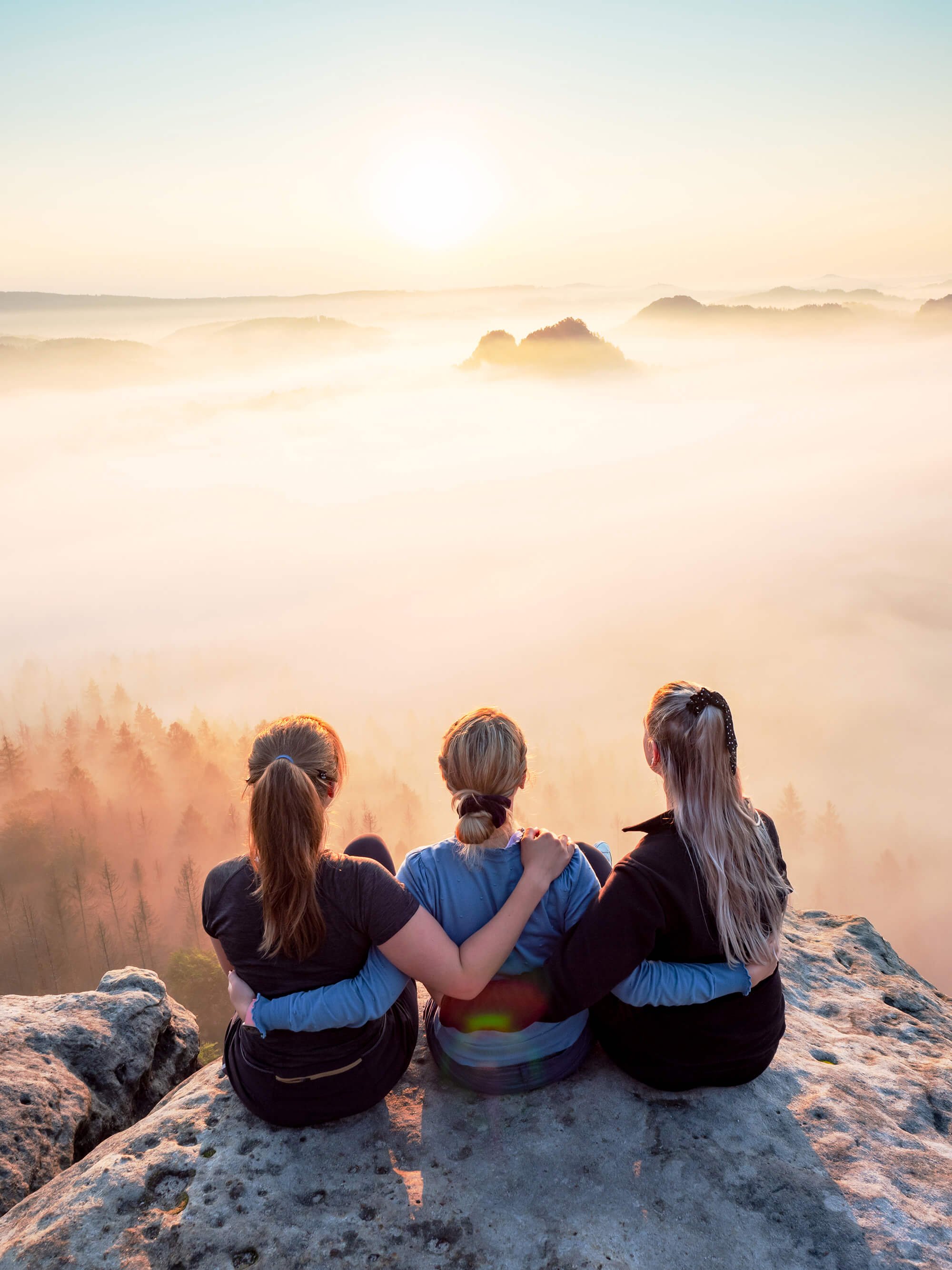 Three women sitting on a rock at sunrise, overlooking a foggy valley with distant hills and trees.