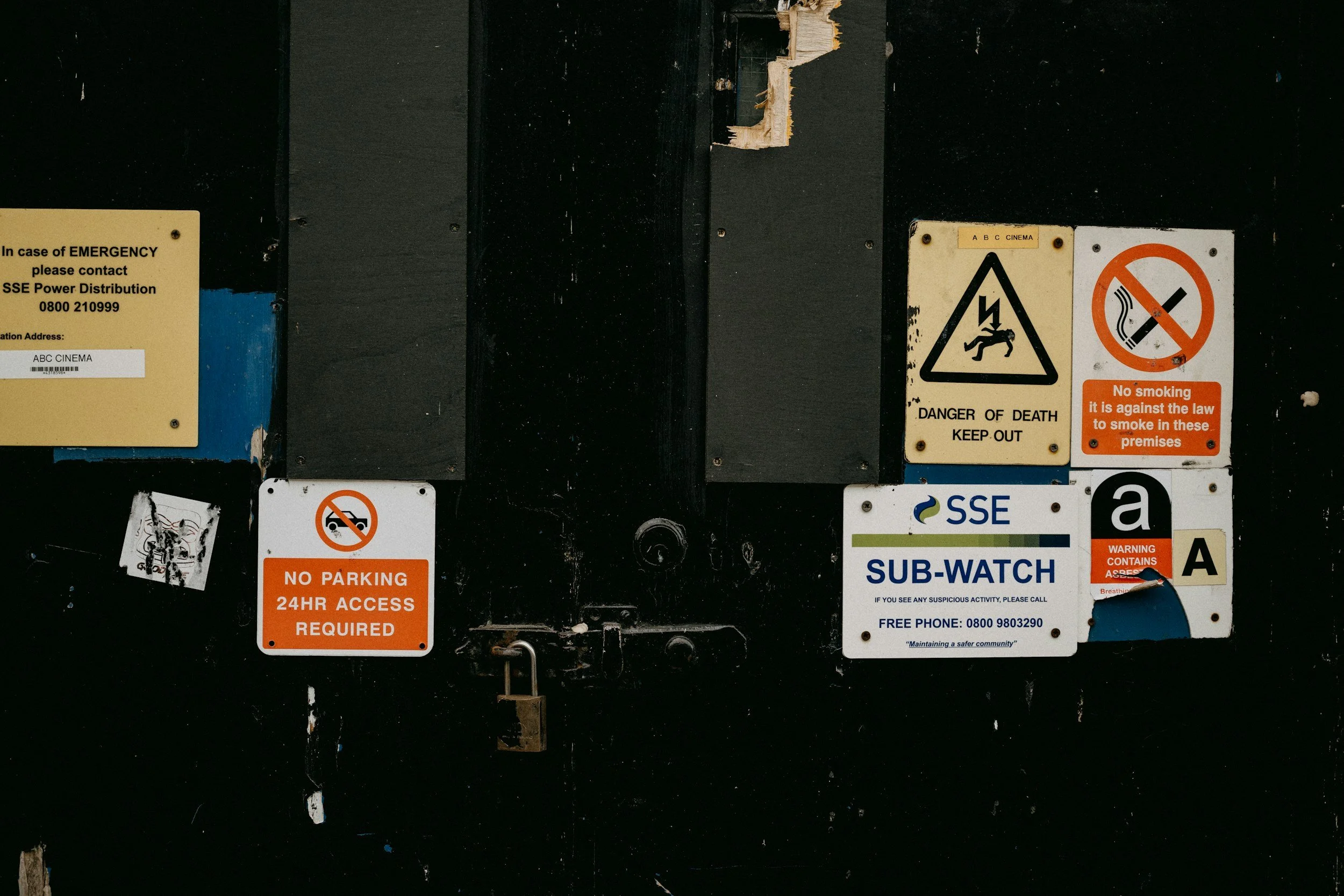 A black metal wall with multiple warning and safety signs, including a yellow emergency contact sign, a no parking sign, a danger of death warning, no smoking sign, and a sub-watch sign from SSE.
