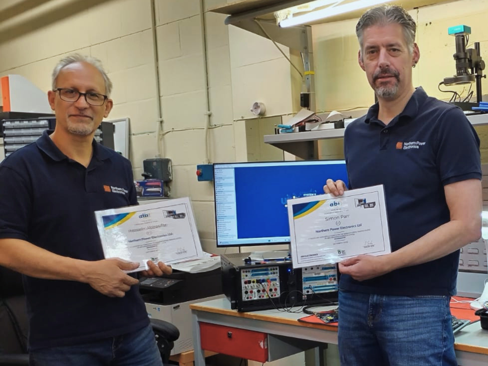 Two men stand in a workshop holding certificates. They are wearing navy blue shirts with the logo of Northern Power Electronics. Behind them is a computer monitor, electronics equipment, workbench, and machinery.
