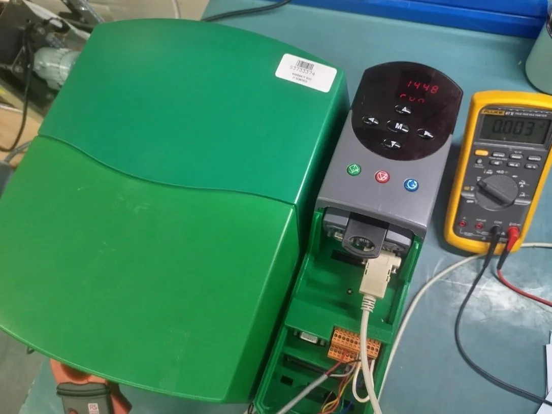 Close-up of a green electronic device with a digital display and control buttons, connected to a multimeter measuring 0.83 volts, on a blue work surface with other tools and wires around.