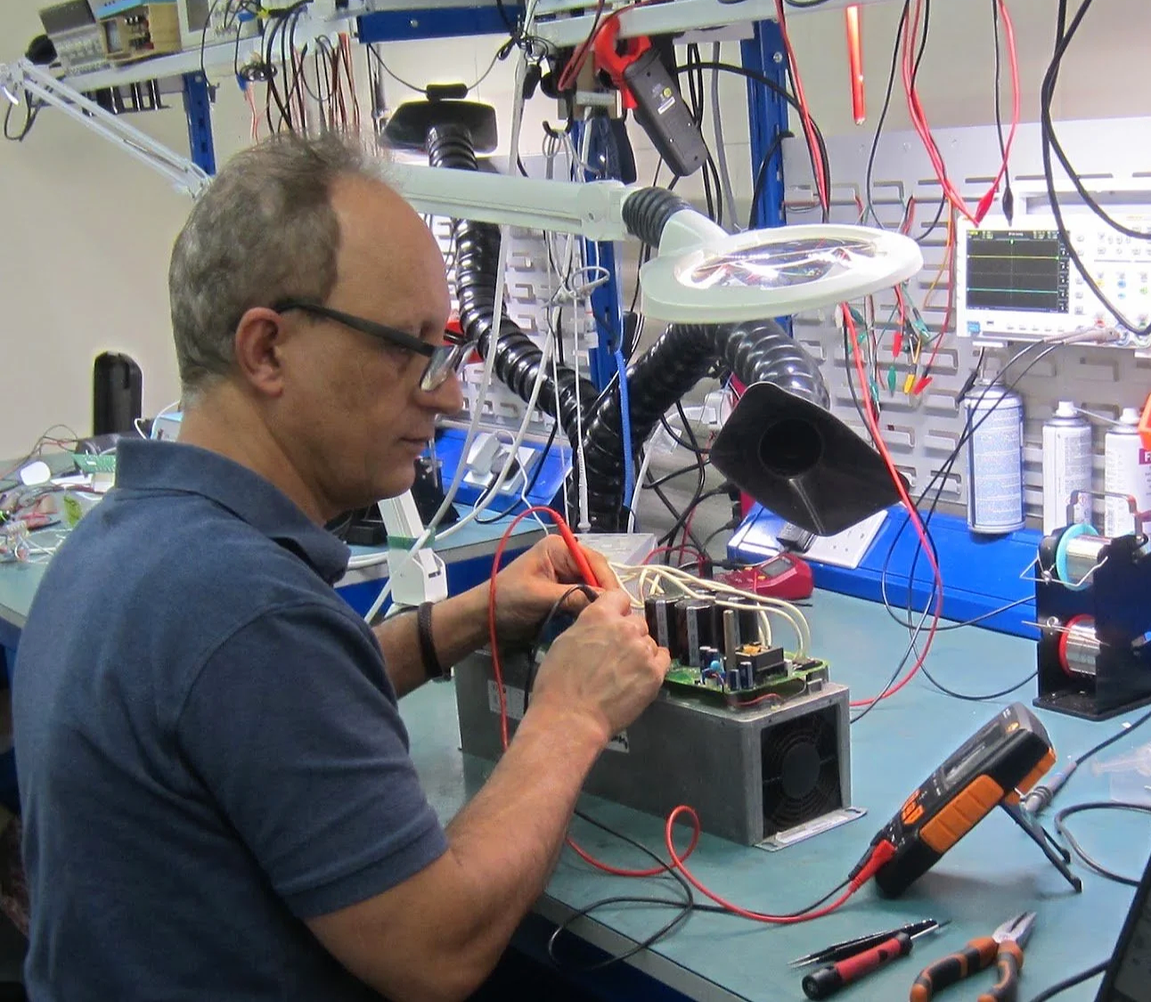 A man working on an electronics project at a cluttered workbench with tools, wires, and electronic equipment.