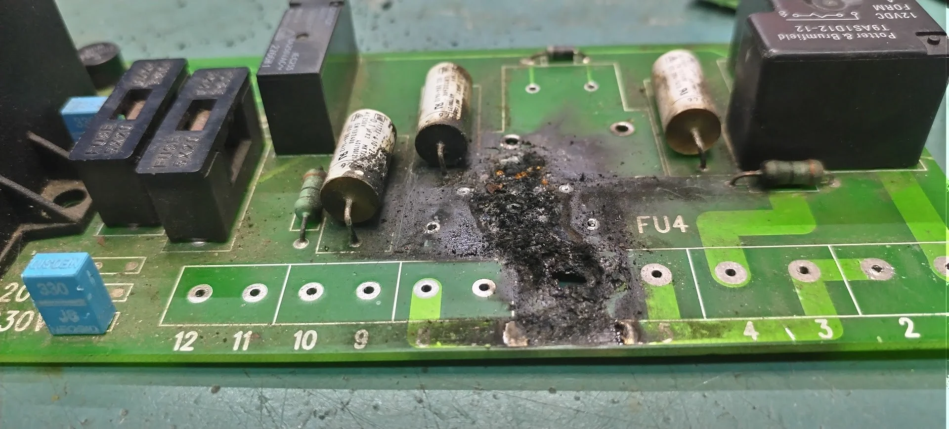 Damaged industrial circuit board with burnt components and blown capacitors awaiting repair at NPE Newcastle"