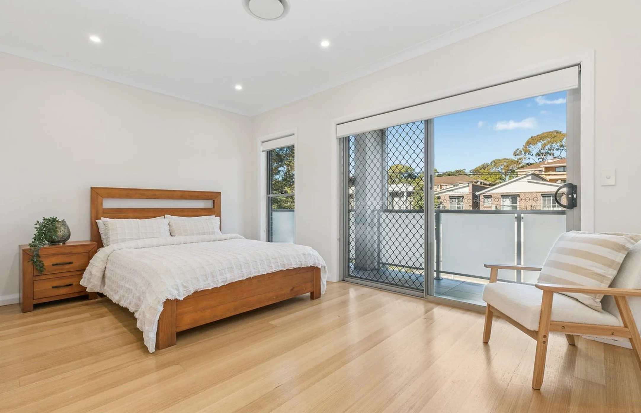 A clean, bright bedroom with a wooden bed, white bedding, a matching wooden nightstand with a plant, a chair with a striped pillow, hardwood floors, and large sliding glass doors leading to a balcony with a view of neighboring houses and blue sky.