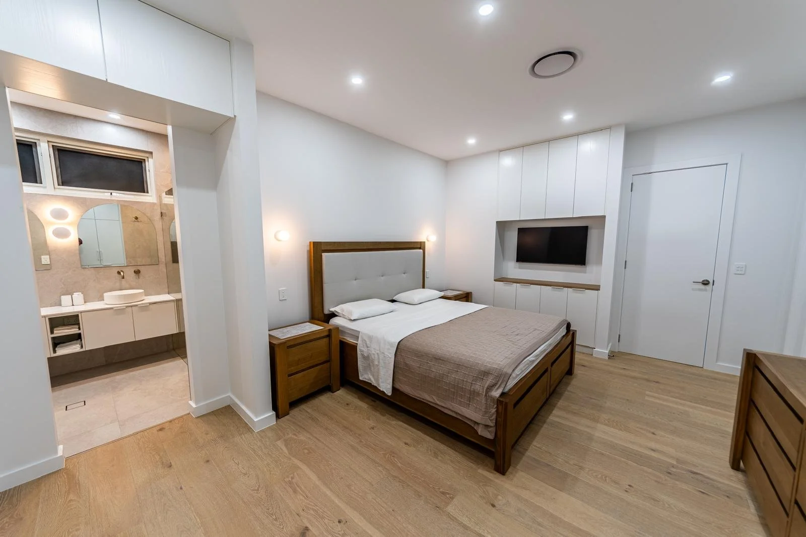 Modern bedroom with wooden furniture, a bed with white and beige bedding, nightstands, a wall-mounted TV, and a visible bathroom with a large mirror and a vessel sink.