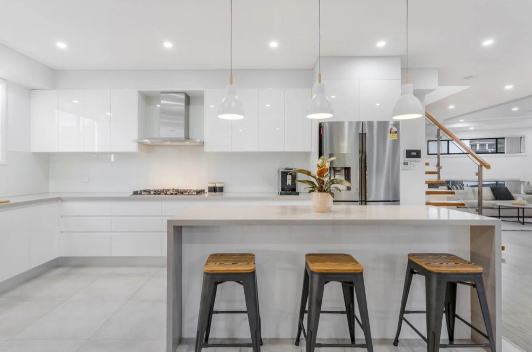 Modern white kitchen with island and three pendant lights, stainless steel refrigerator, plant on island, and view of living room and staircase