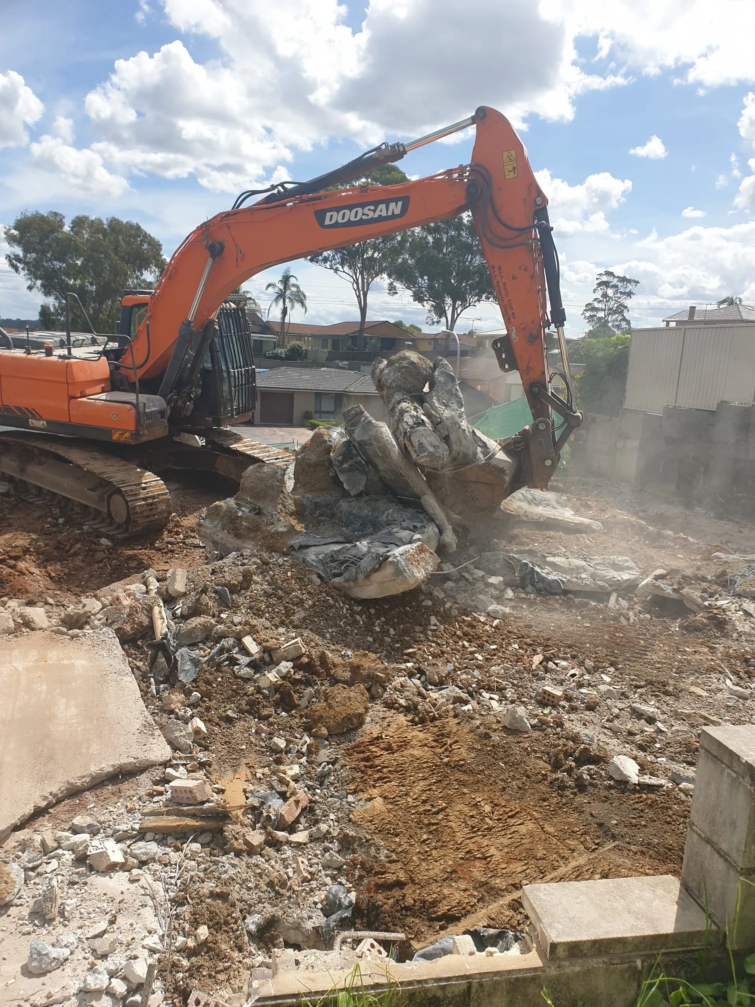 An orange Doosan excavator demolishing concrete and rocks on a construction site under a partly cloudy sky.