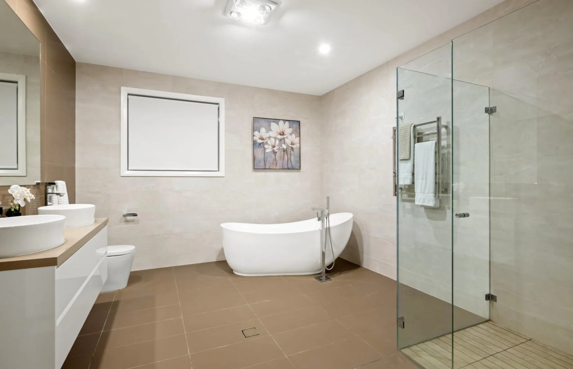 Modern bathroom with bathtub, glass-enclosed shower, white dual sinks, toilet, beige tiles, a framed flower painting, and a window.