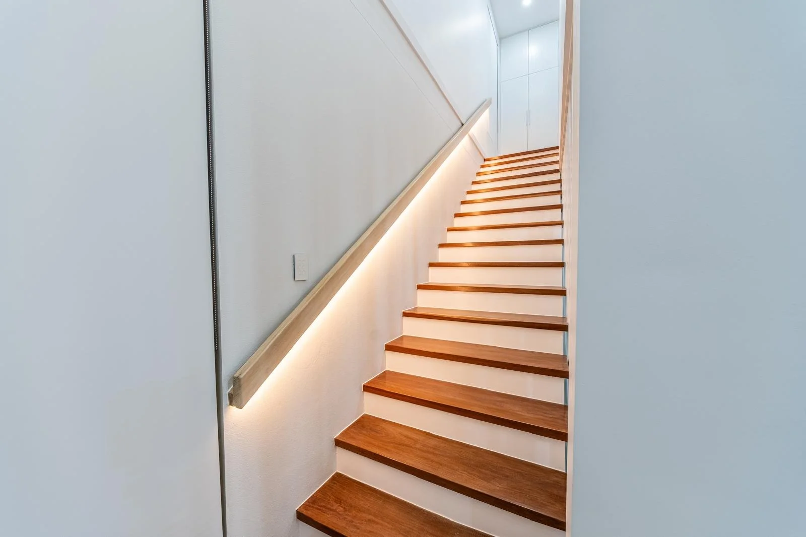 Modern wooden staircase with a built-in wall light and a sleek handrail, ascending between white walls to an upper level.