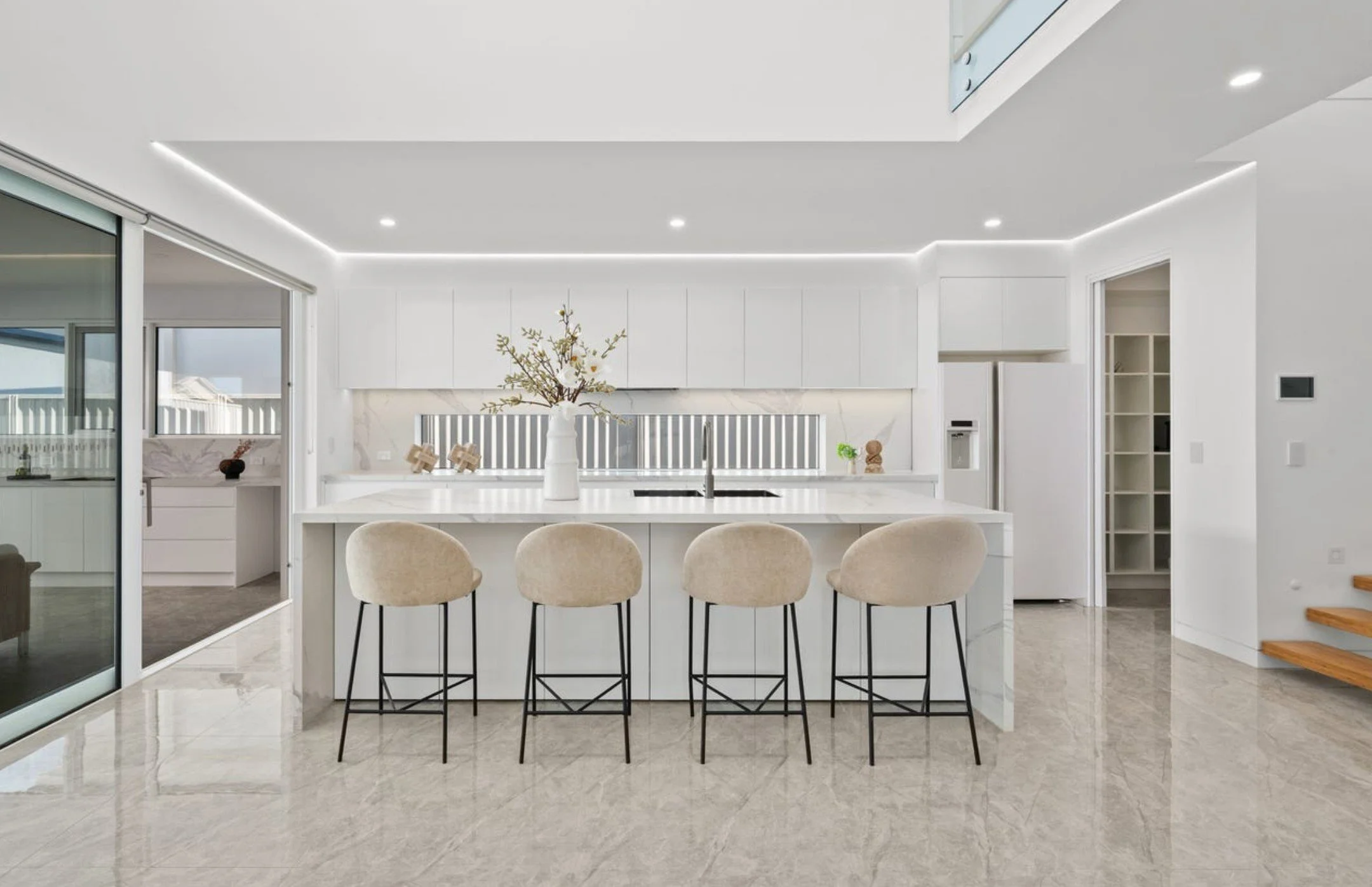 Modern white kitchen with marble flooring, four beige barstools at a white island, and a large vase with branches as decoration.