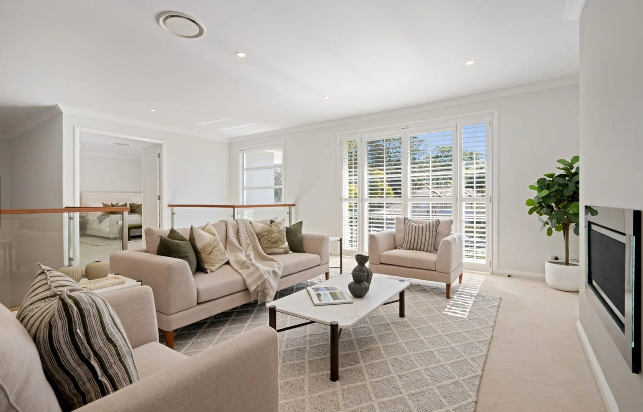Bright living room with white walls, large window with blinds, beige sofas with pillows, a white coffee table, potted plant, and wall-mounted TV.