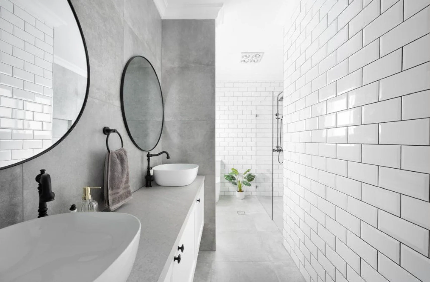 Modern bathroom with gray concrete wall, white vanity with two vessel sinks, black fixtures, large oval mirrors, white subway tile walls, and a walk-in shower with a glass partition and a plant in the corner.