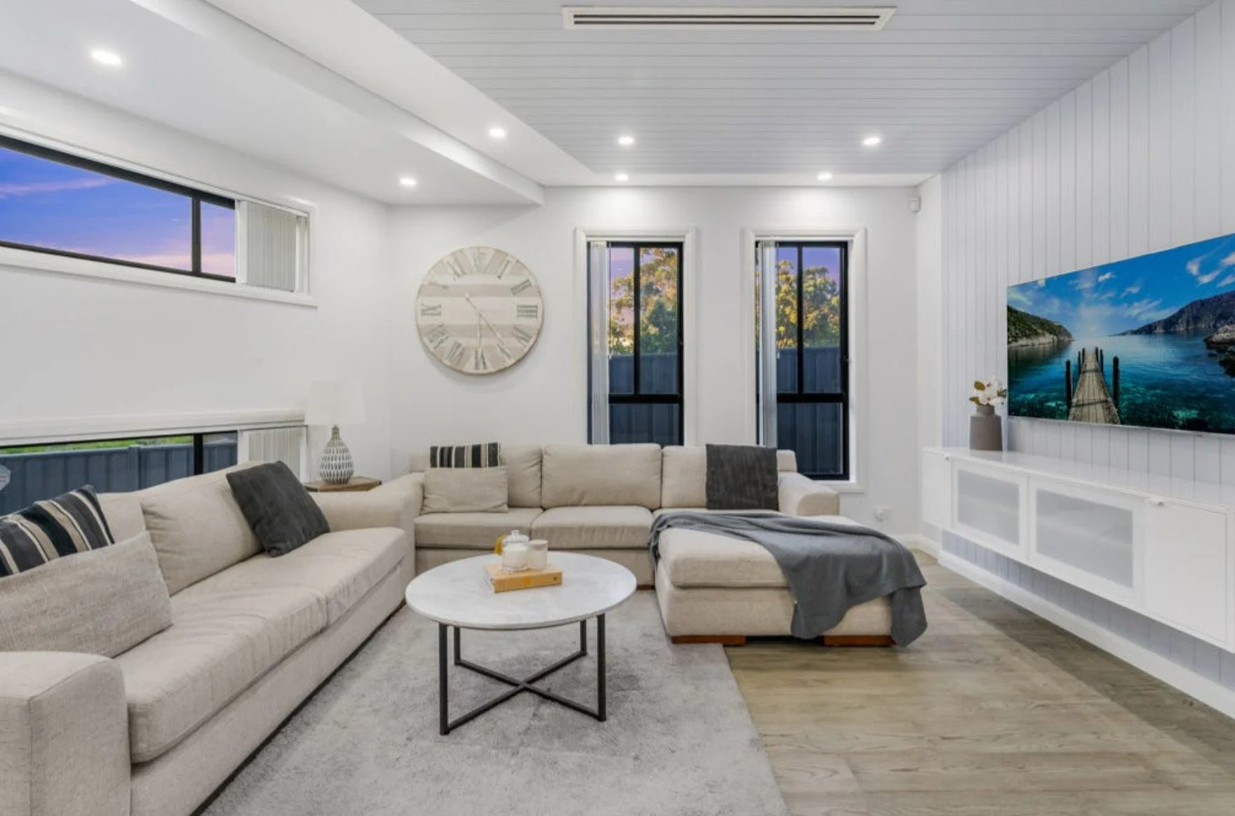 Living room with white walls, beige sectional sofa with black and beige pillows, round white coffee table, large wall clock, windows with outdoor view, flat-screen TV, gray throw blanket, lamp, decor items