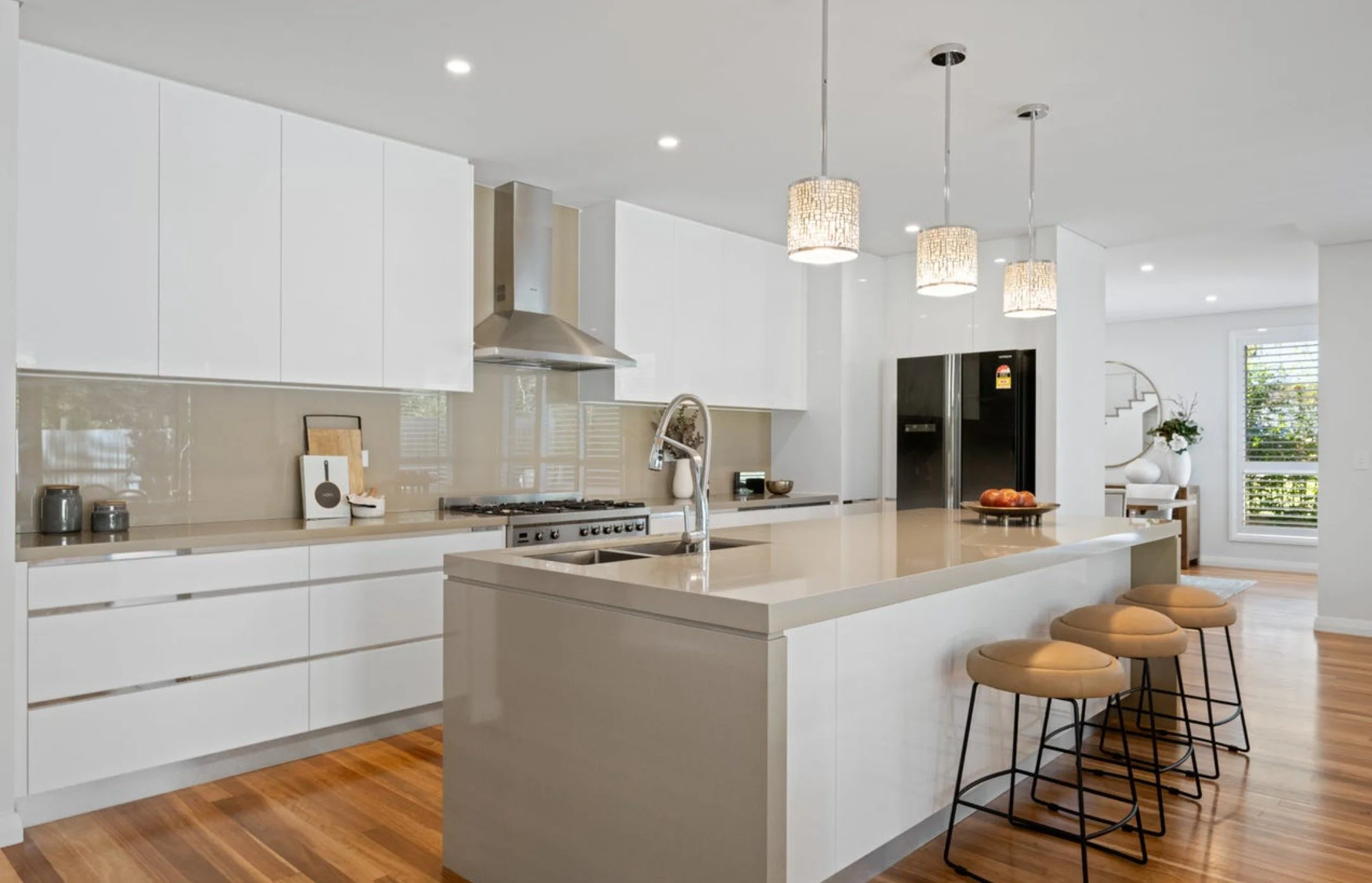Modern kitchen with white cabinets, beige countertops, and a large island with seating. Stainless steel appliances, pendant lights, and hardwood floors. A refrigerator and a window with blinds are visible.