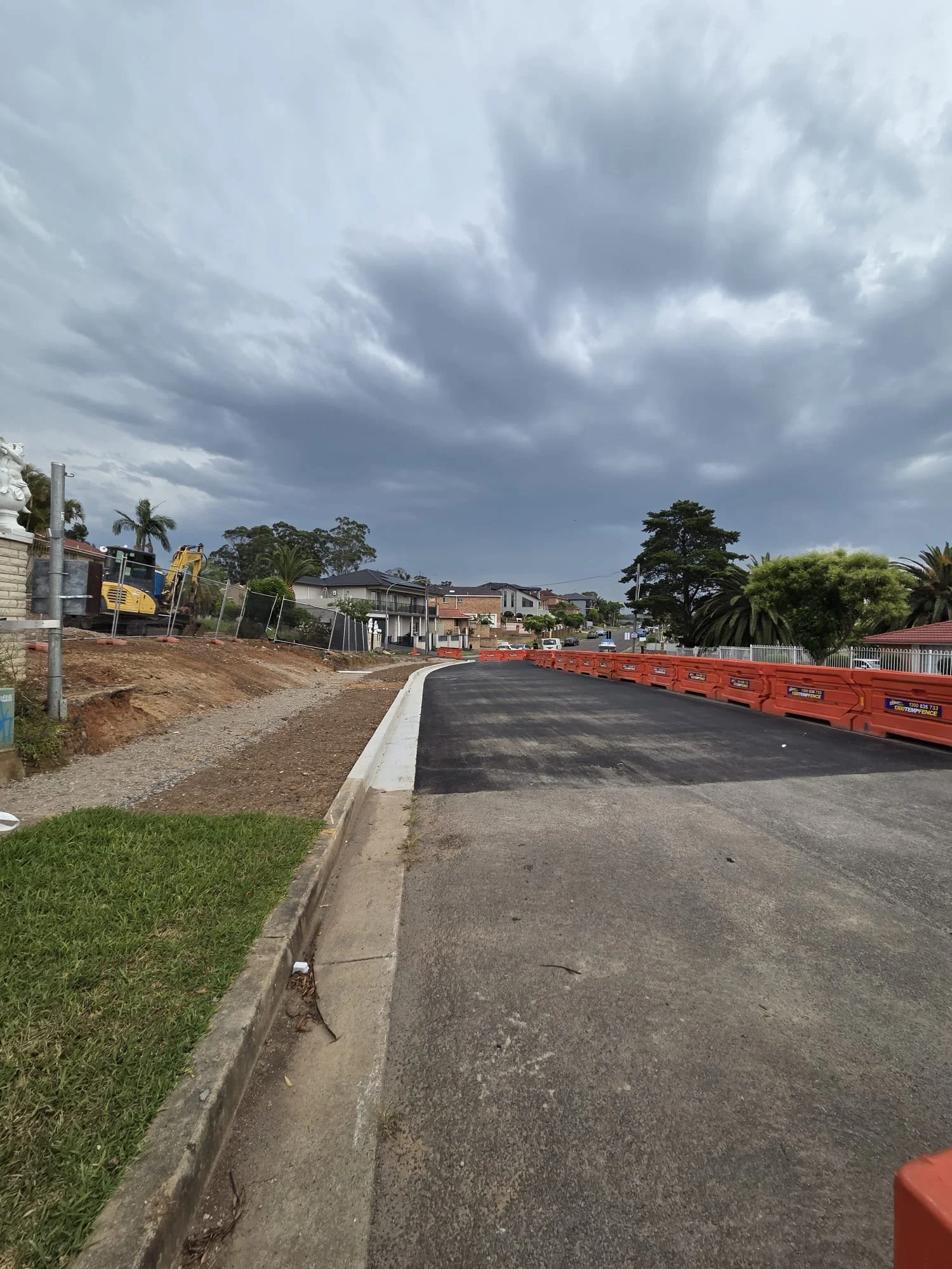 A street under construction with a sidewalk on the left and orange safety barriers on the right. The road appears to be newly paved, and there are homes, trees, and a cloudy sky in the background.