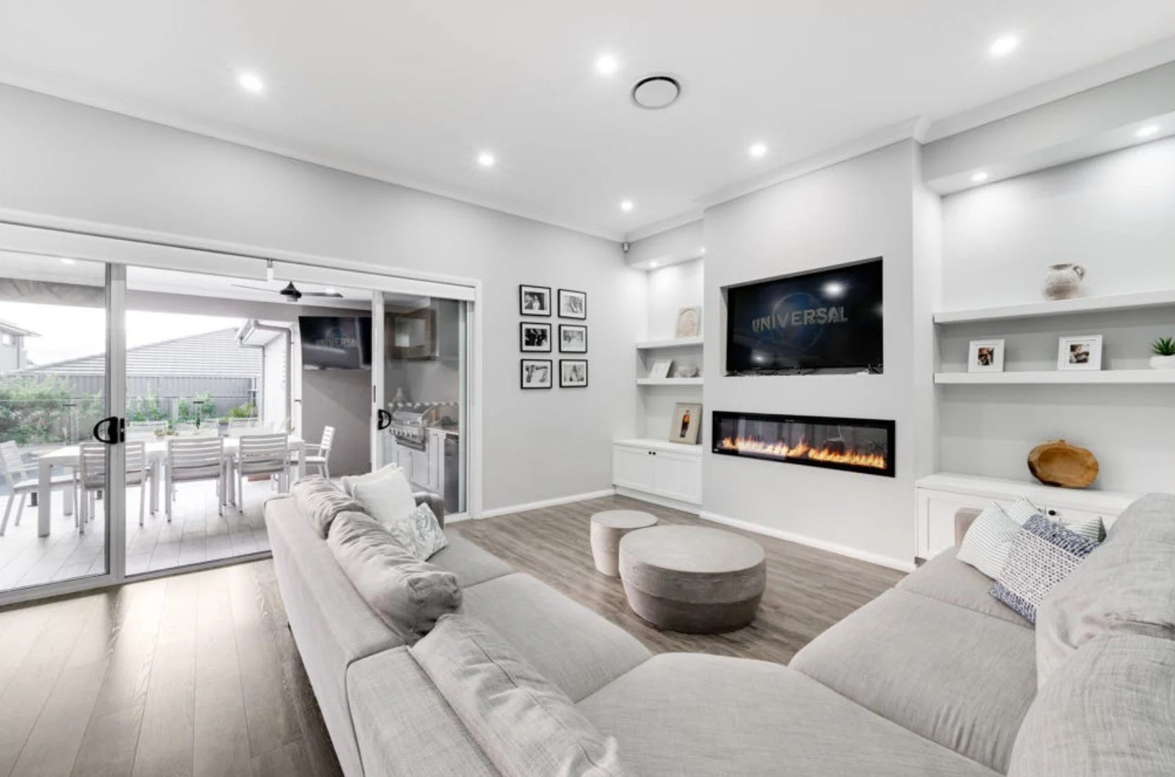 Living room with white walls, ceiling lights, a large beige sofa, a gray round coffee table, a wall-mounted TV displaying 'Universal', and a built-in fireplace below the TV. Sliding glass doors lead to an outdoor patio with a dining table and chairs.