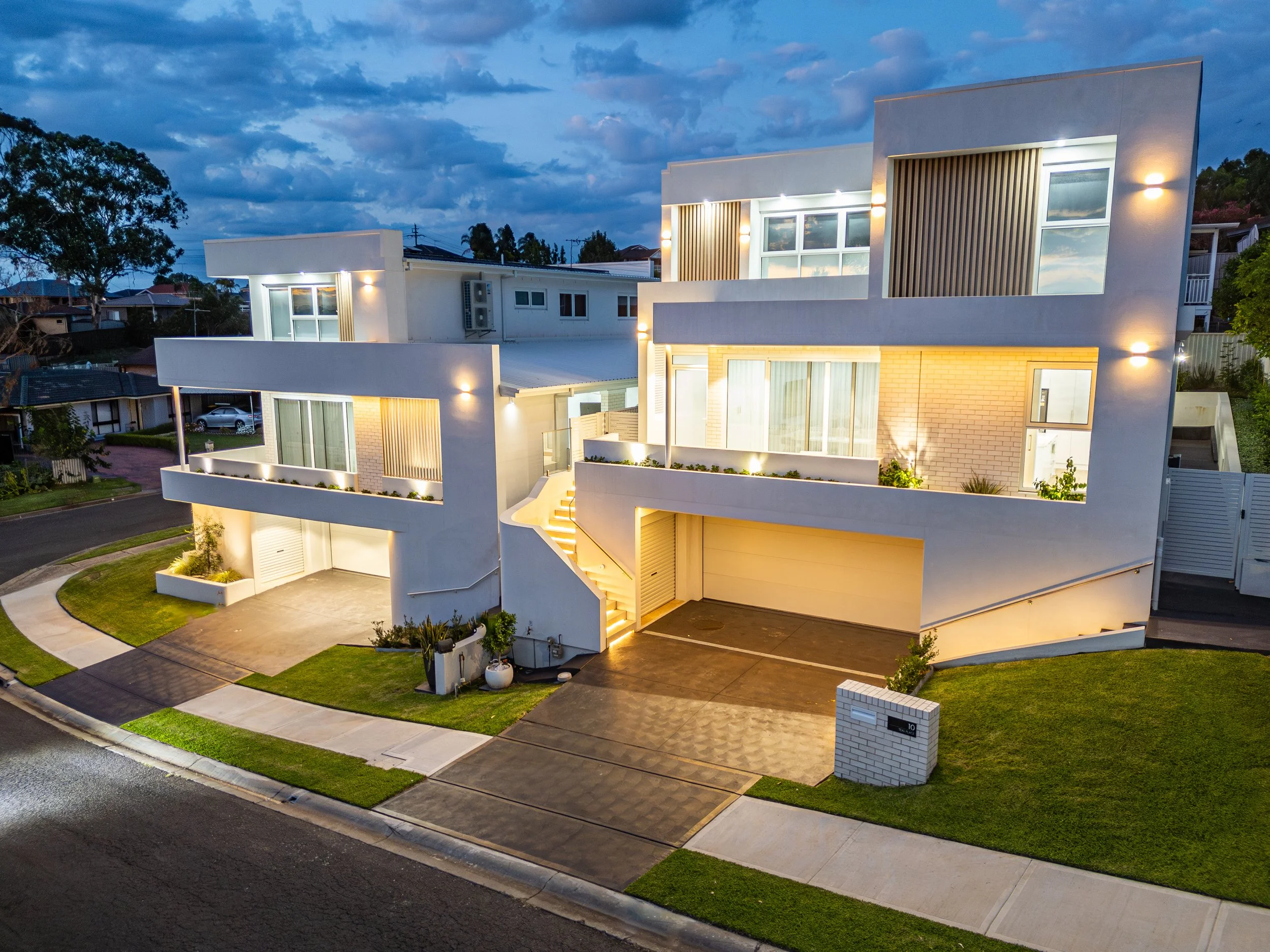 Modern multi-story house illuminated with exterior lights during dusk, featuring large windows, balconies, a garage, and landscaped yard.