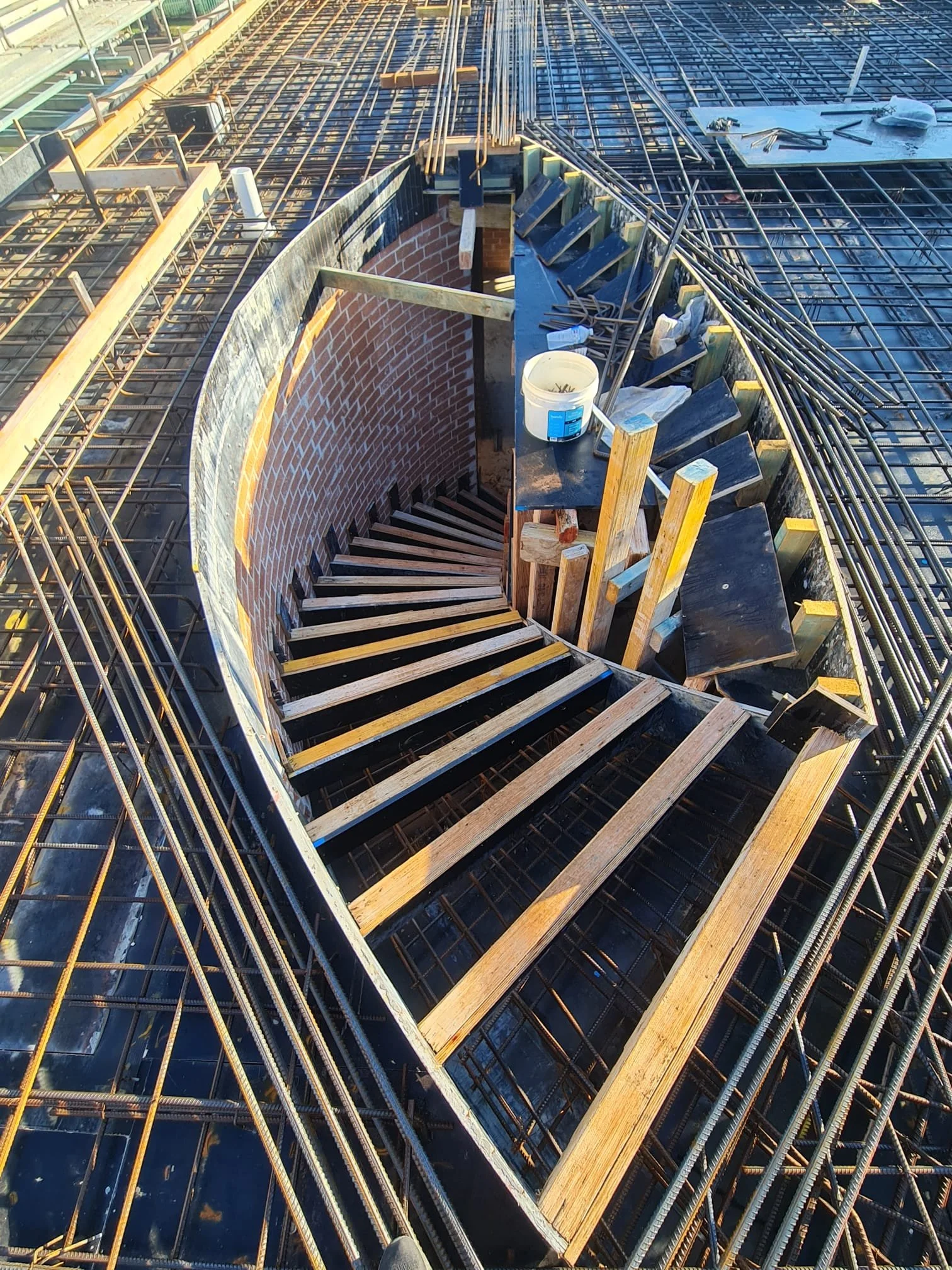 Construction site showing a spiral staircase under construction with recessed brick wall, wooden steps, and metal framework around the opening.