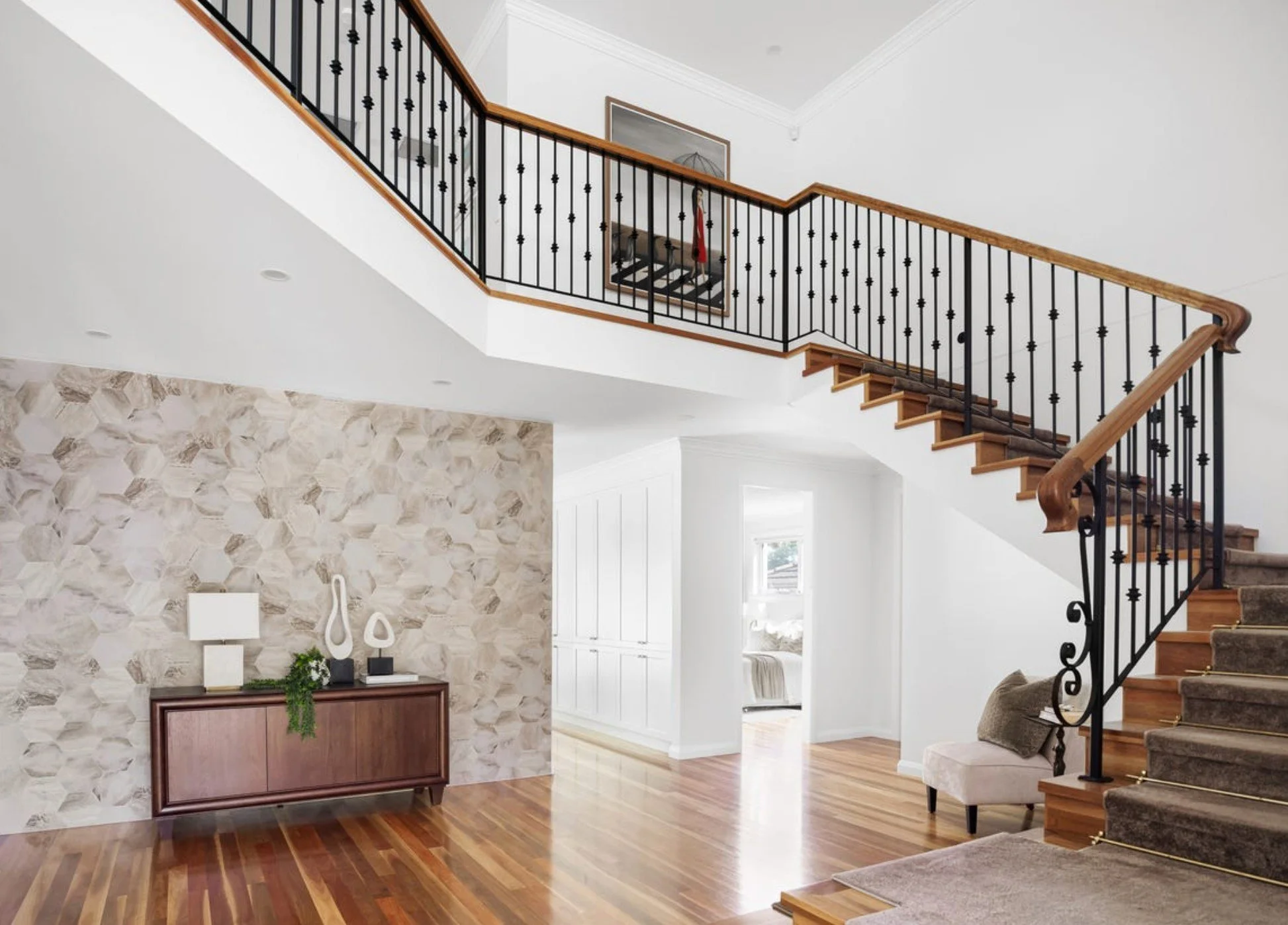 Bright interior of a modern home with a staircase featuring black metal balusters and wooden handrail. The living area has a decorative stone accent wall, a wooden sideboard with modern sculptures and a lamp, and a beige upholstered chair. There is n