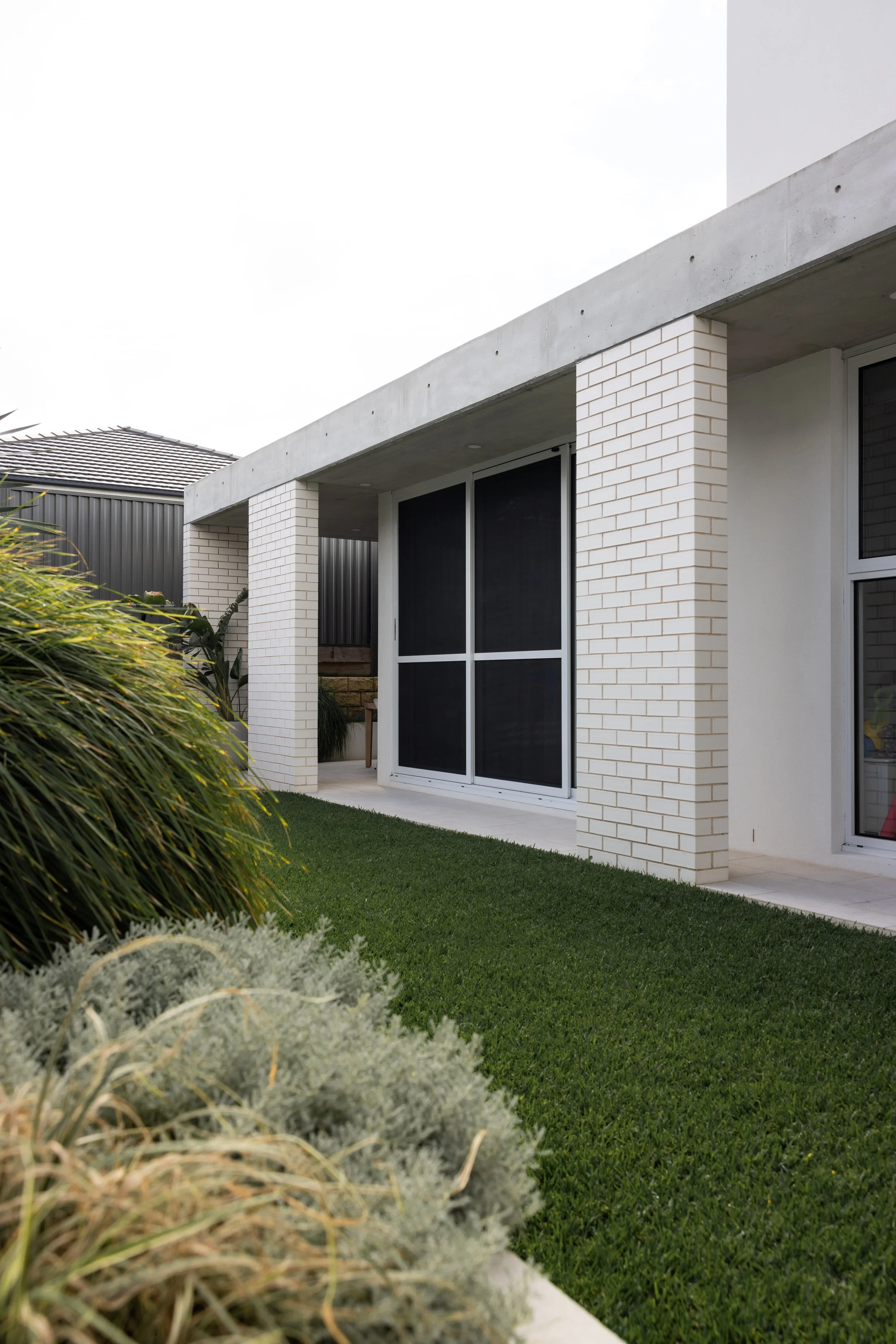 Modern house with white brick pillars, large sliding glass door with black screen, and neatly maintained lawn and garden in the foreground.