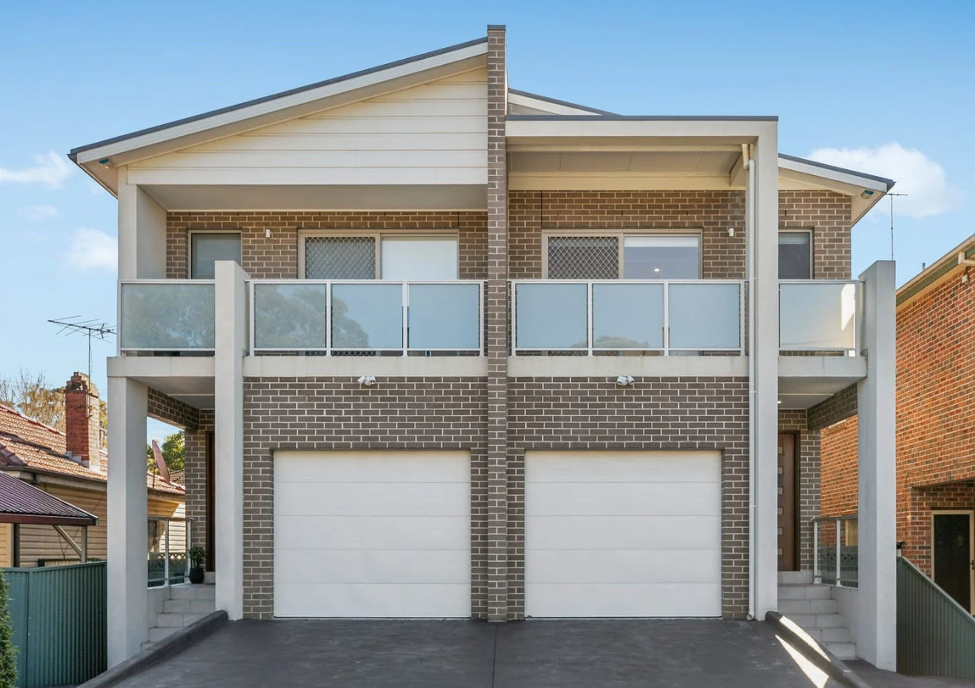 Modern two-story brick residential building with two garage doors on the ground level, balconies with glass railings on the upper level, and stairs on both sides leading to the entrances.