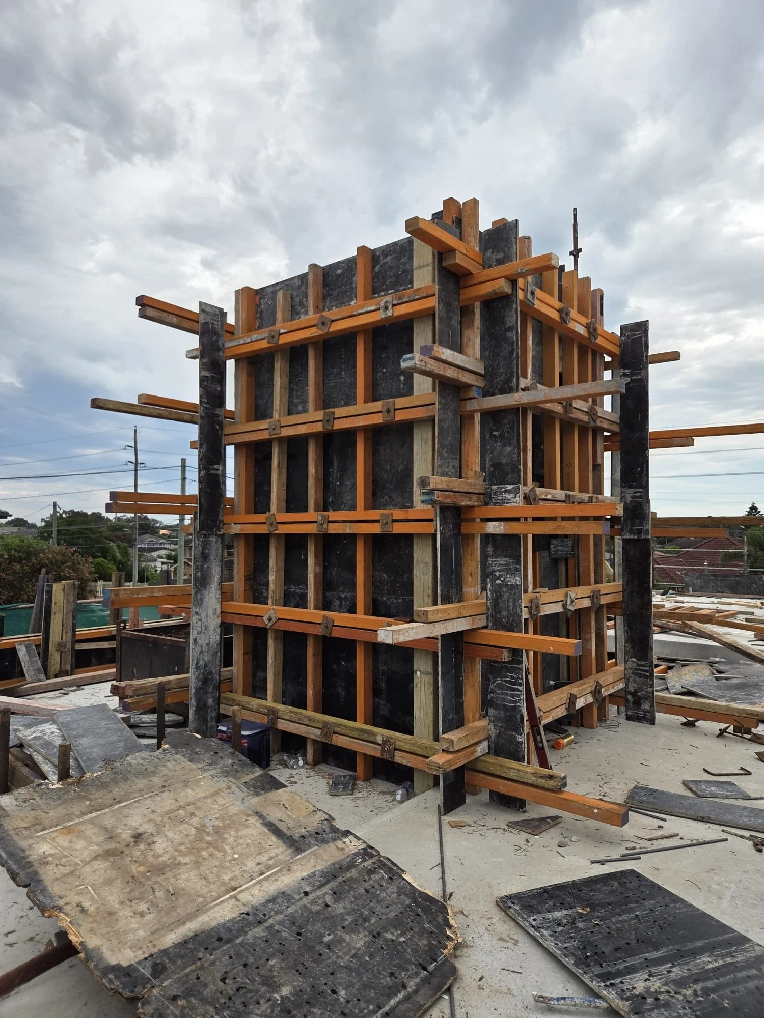 Construction site with wooden framework and black formwork for a concrete wall, with cloudy sky overhead.