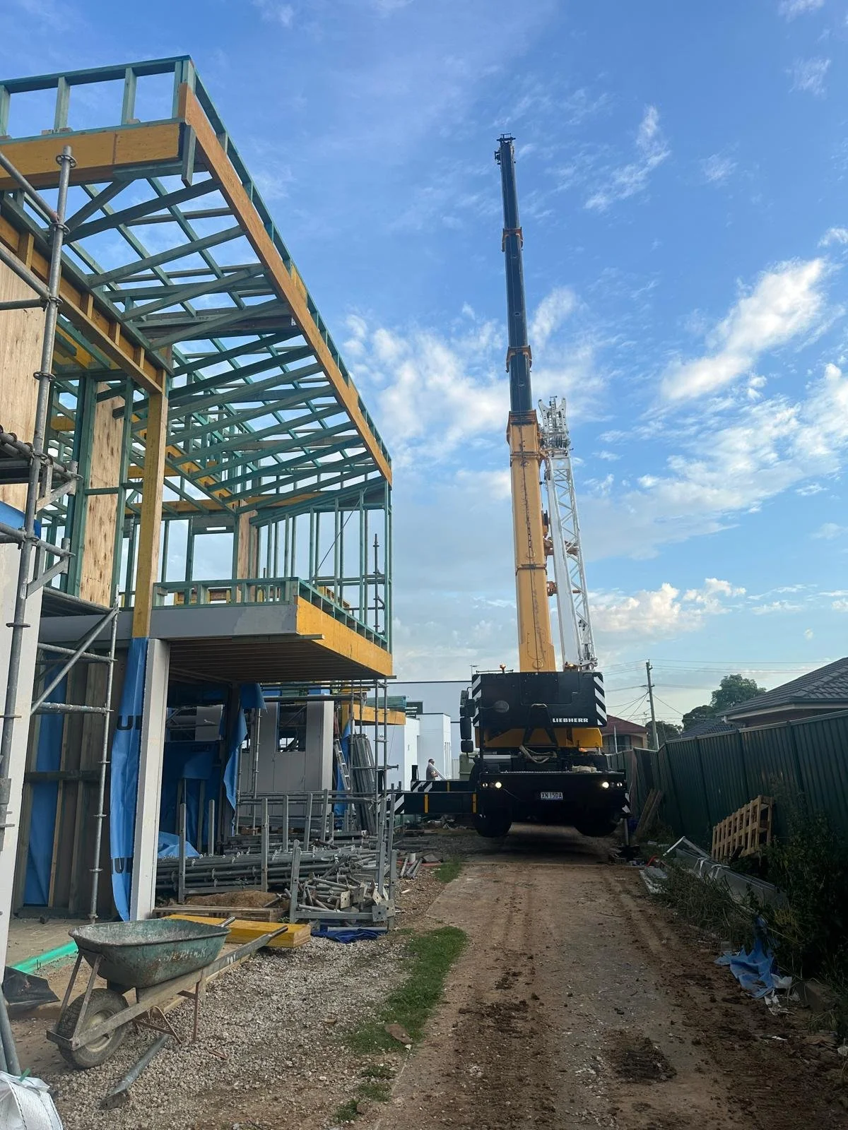 Construction site with a large crane lifting materials near a partially built multi-story building, surrounded by scaffolding and construction tools under a blue sky with scattered clouds.