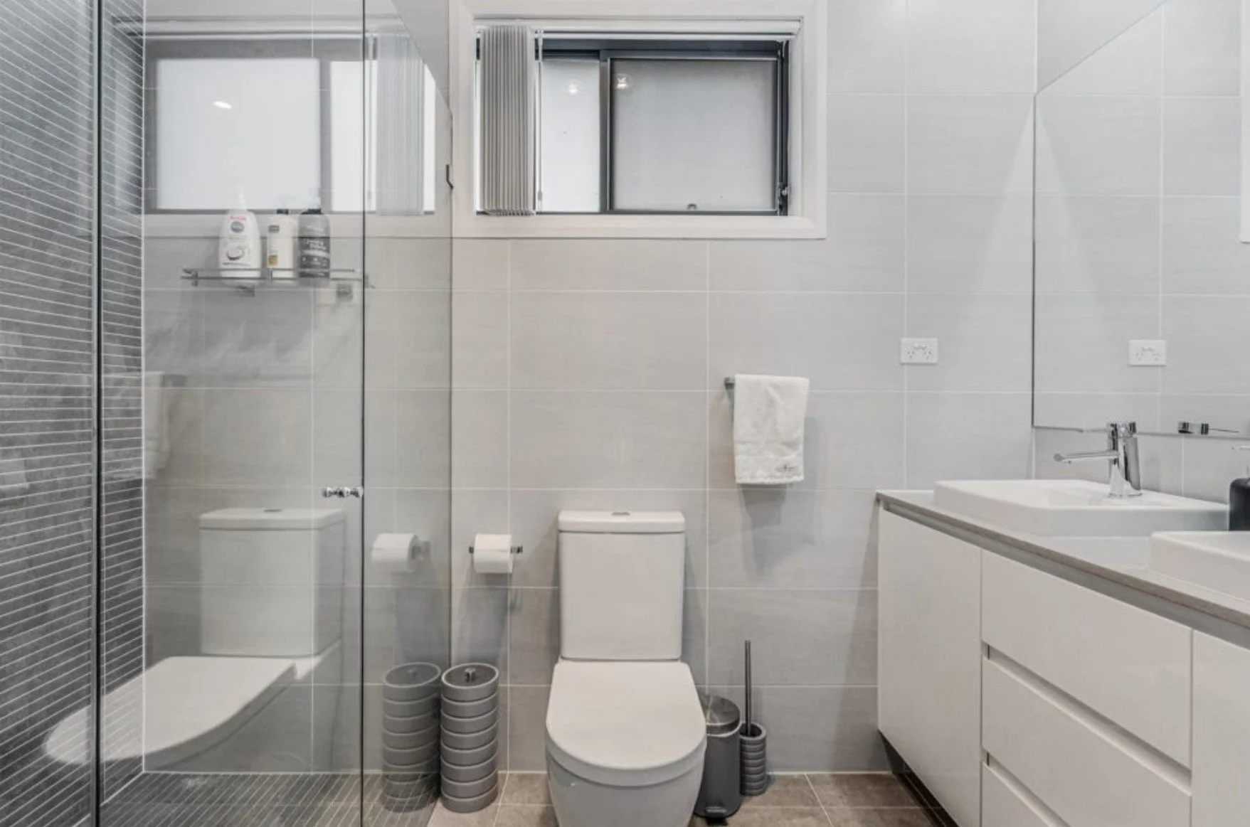 Modern bathroom with white tiles, a toilet, a glass shower enclosure, a vanity with two sinks, a mirror, and a small window.