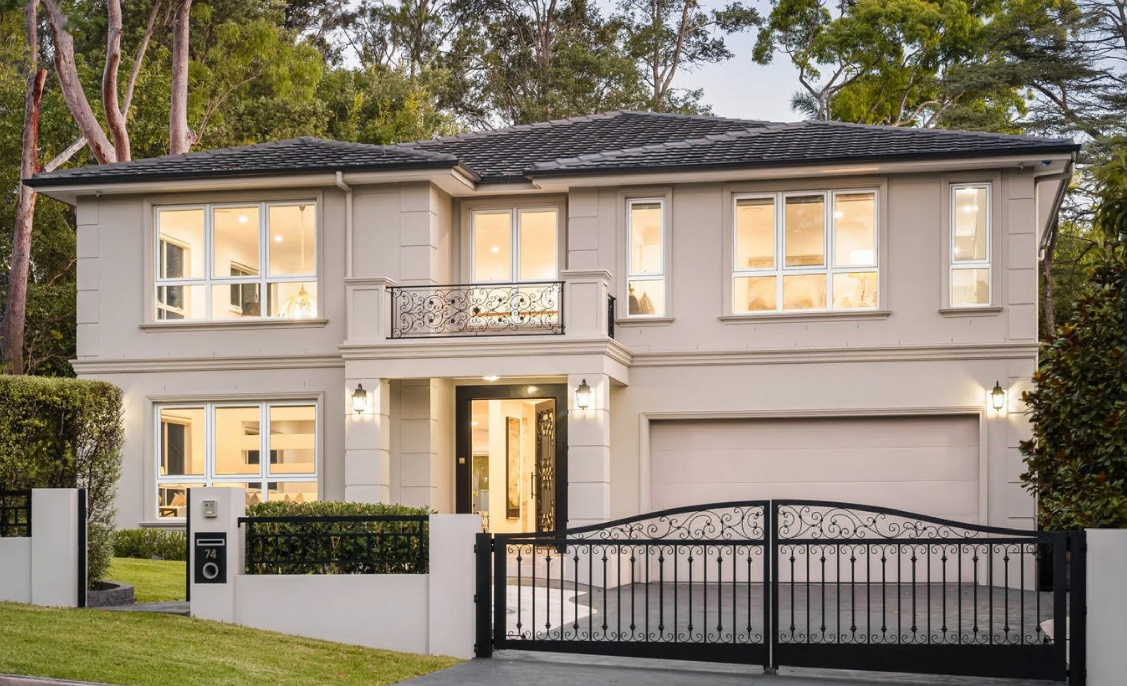 A two-story white house with decorative black wrought iron gate and fence, large illuminated front porch, multiple windows, and a lush green lawn, surrounded by tall trees.