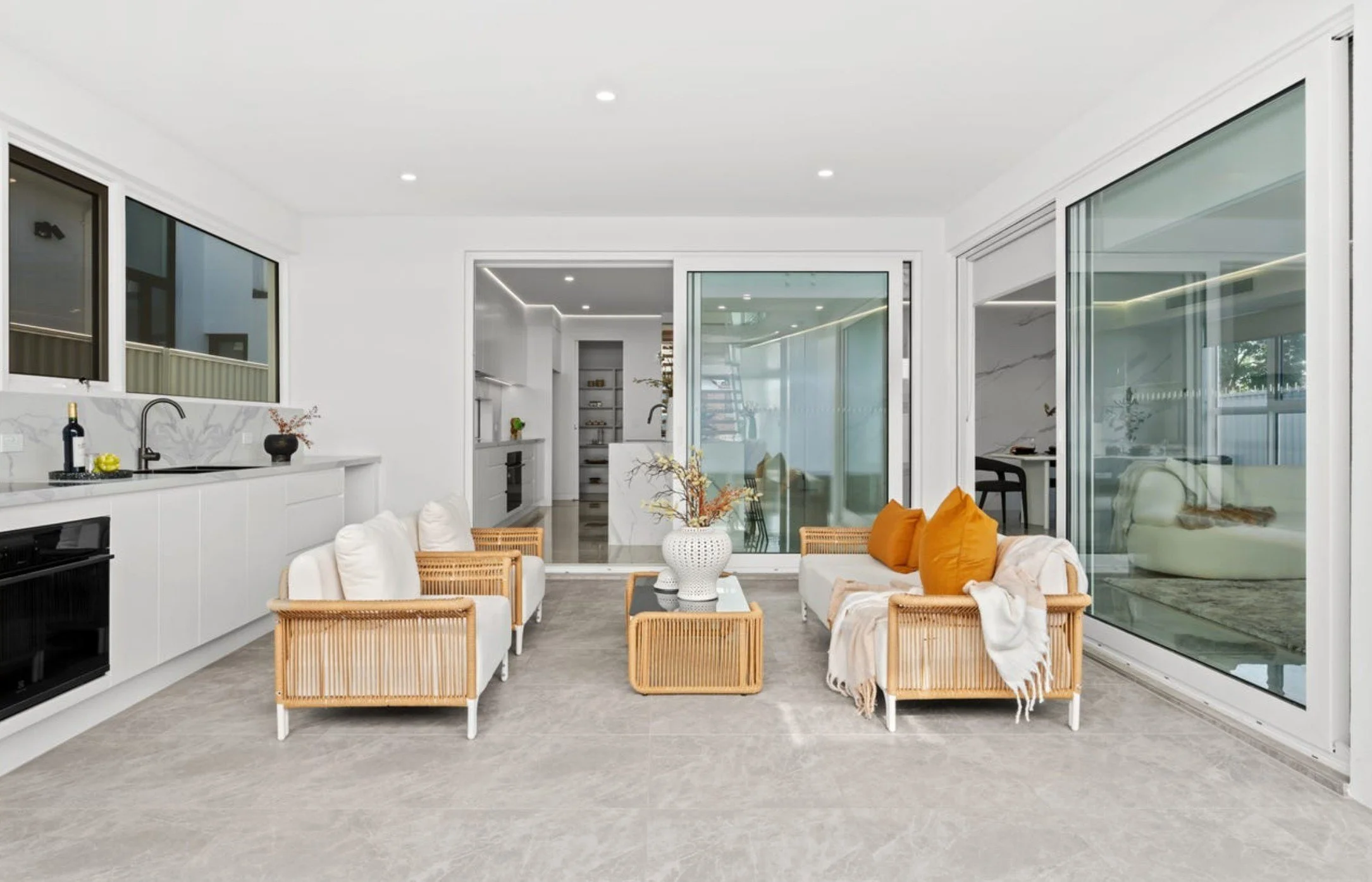Bright living room with white walls, large windows, and glass sliding doors; two white sofas with pillows and throws, rattan furniture, a white vase with flowers on a glass coffee table, and a view into a kitchen and another room.