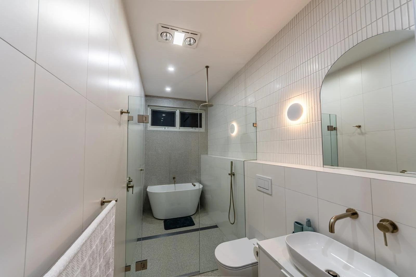 Modern bathroom with white tiled walls, a glass shower partition, a freestanding bathtub, and a toilet. A large mirror reflects circular wall lights, and a small window is visible near the ceiling. There is a basin with a wall-mounted faucet and some