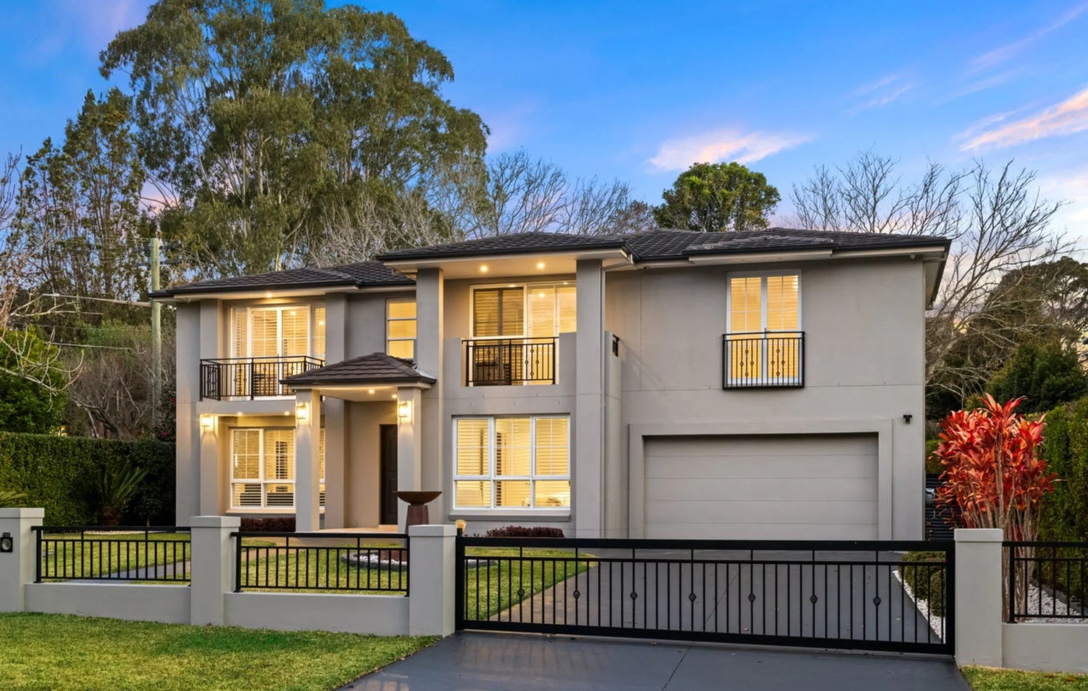 A modern two-story house with a white exterior, black roof, and a garage, illuminated by exterior lights, with a gated front yard, green lawn, and trees in the background during dusk.