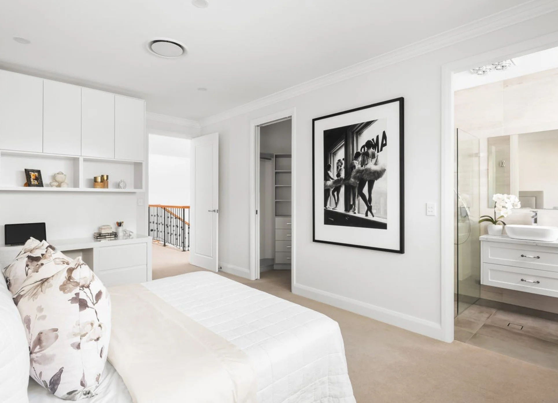 Bright, white bedroom with bed, floral pillows, framed black and white photo on wall, open closet, and bathroom with white vanity and flower